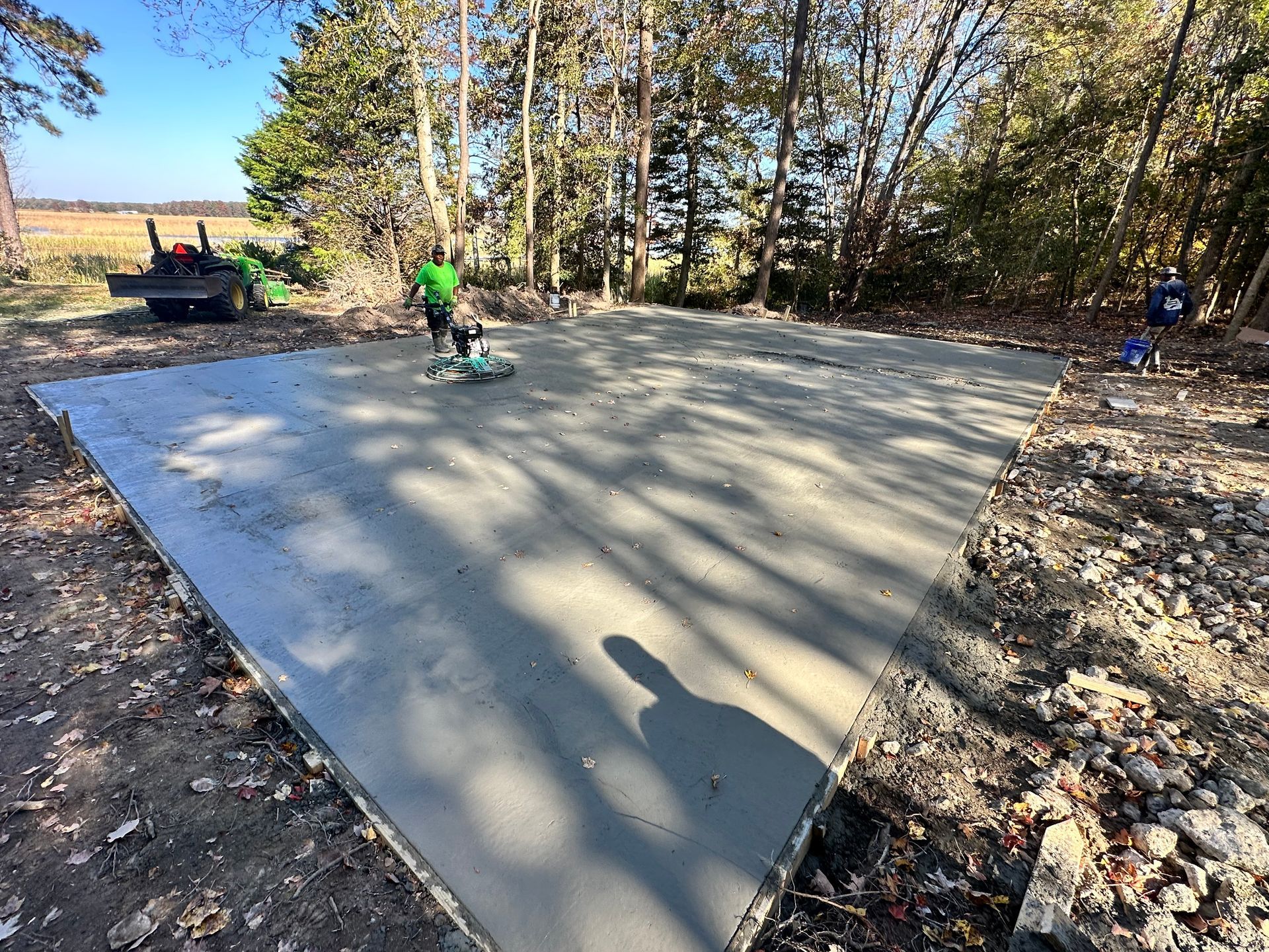 Concrete slab being smoothed by a worker in green, outdoor setting with trees and tractor.