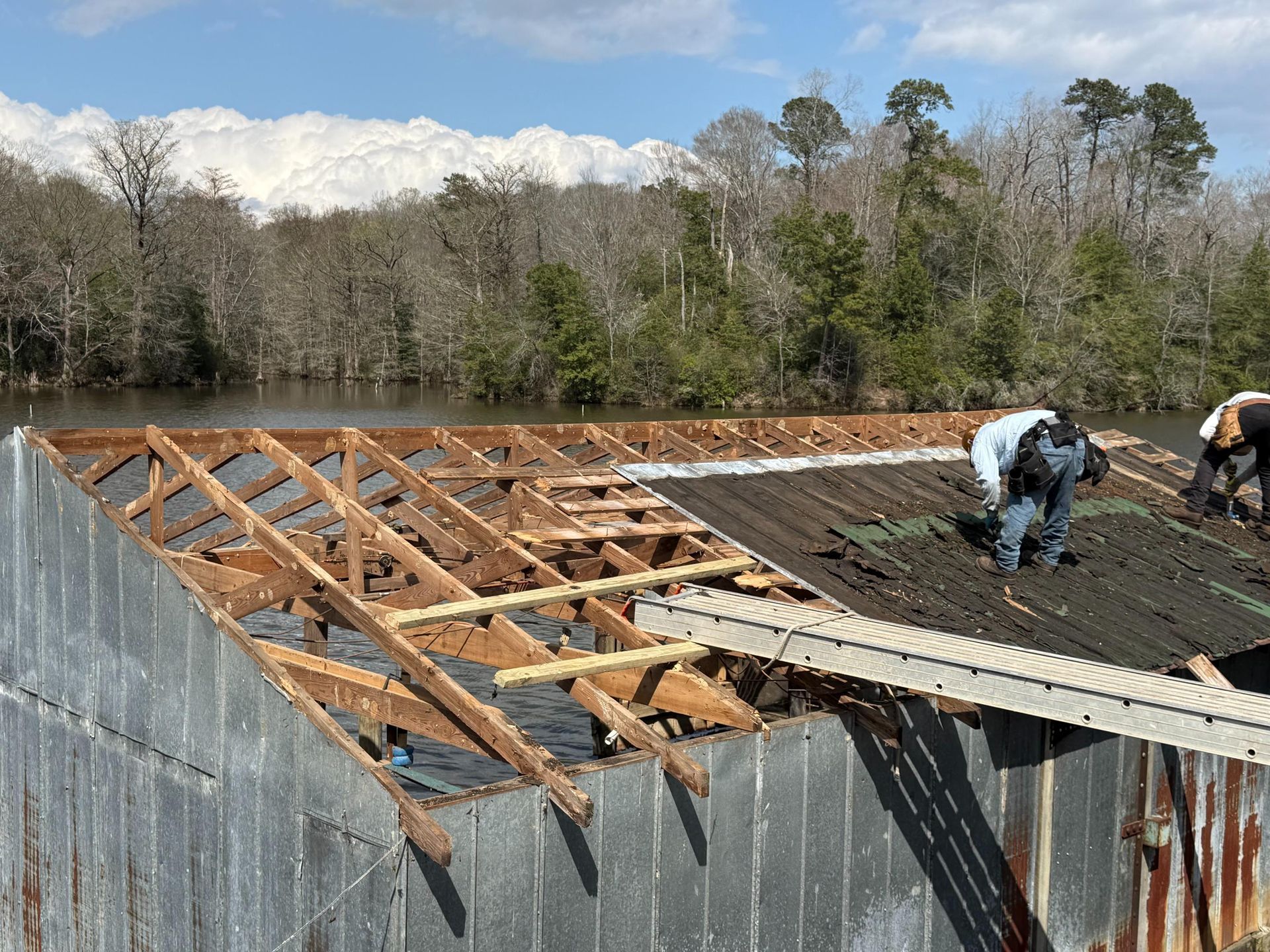 Workers dismantle a weathered metal-roofed structure near a lake and treeline.