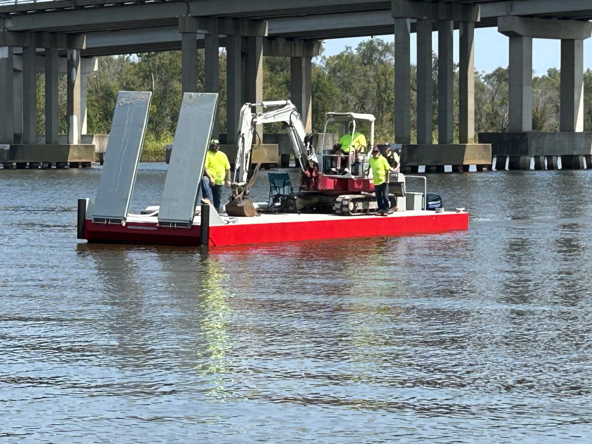 Construction barge on water with excavator, under bridge. Workers in yellow vests. Red barge.