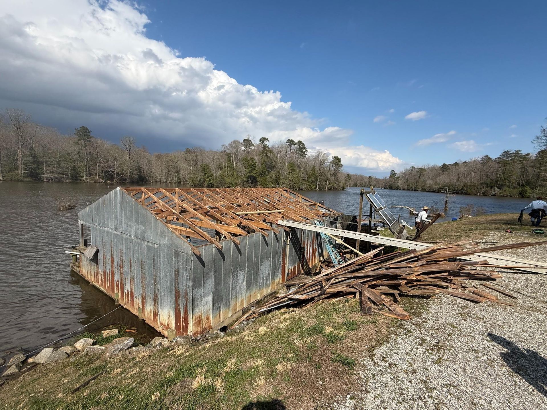 A partially demolished metal shed sits on the bank of a river, with debris scattered on the ground near workers.