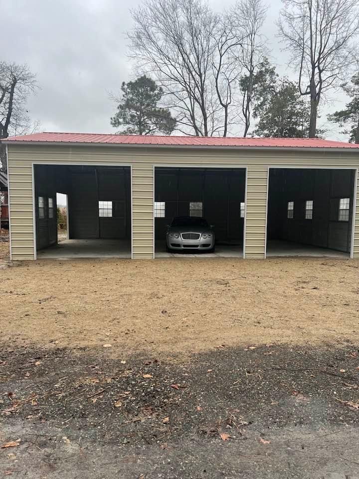 Three-bay metal garage with a red roof, a car parked in the middle bay, on a dirt lot.