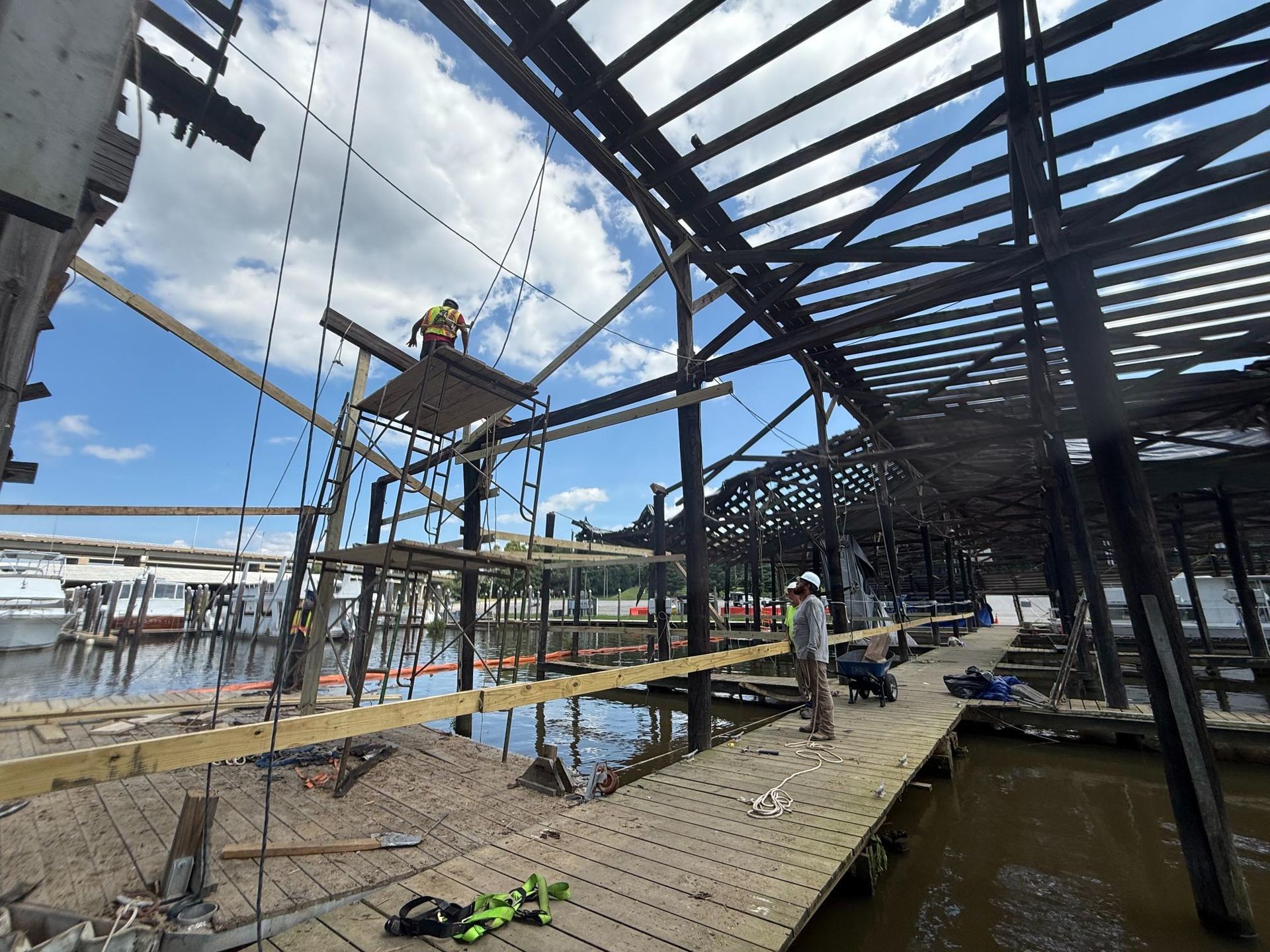 Construction workers rebuilding a dock, with scaffolding and wooden beams on water, under a blue sky.