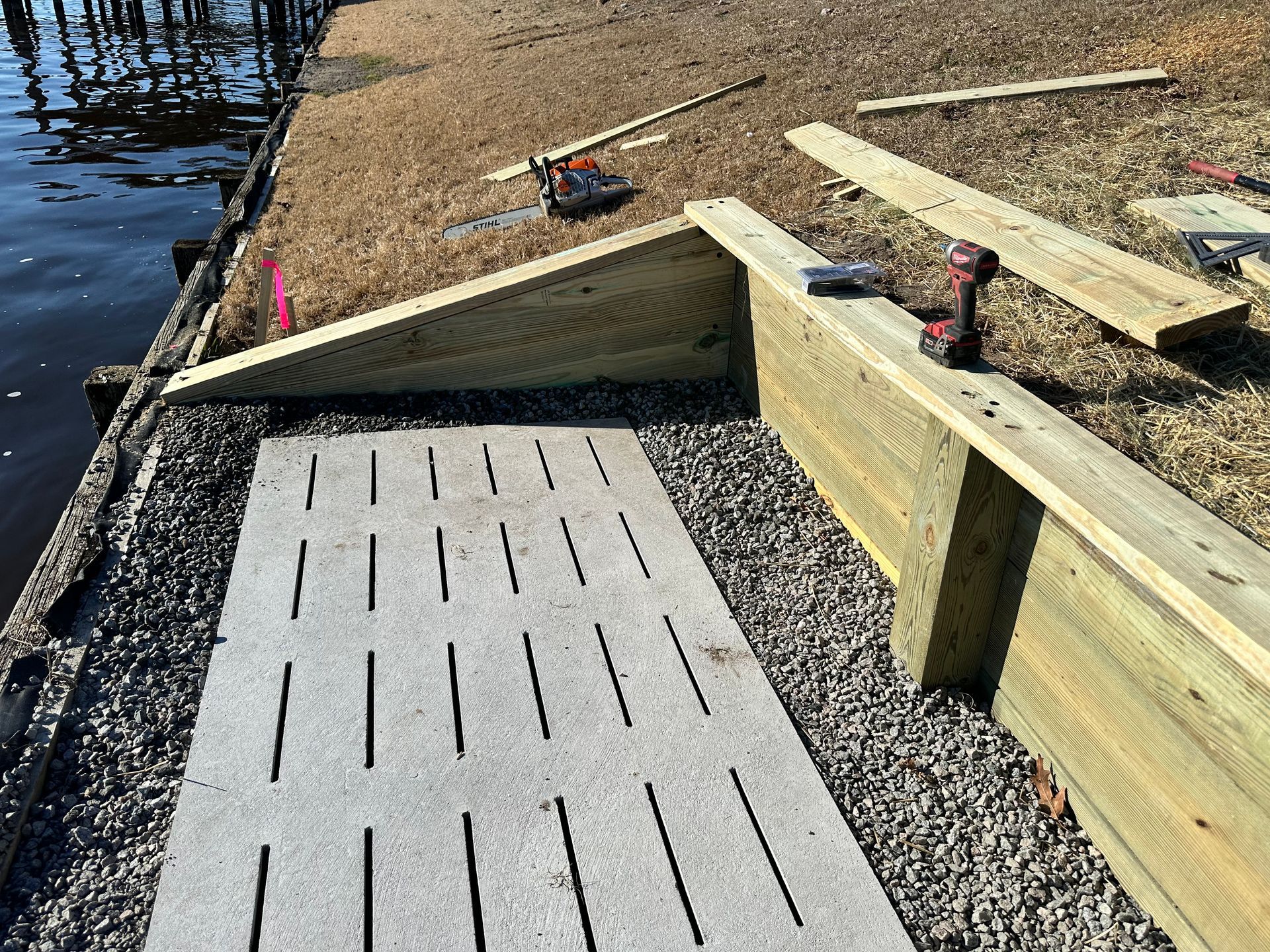 Construction of a wooden dock with concrete pathway, gravel, and water in background.