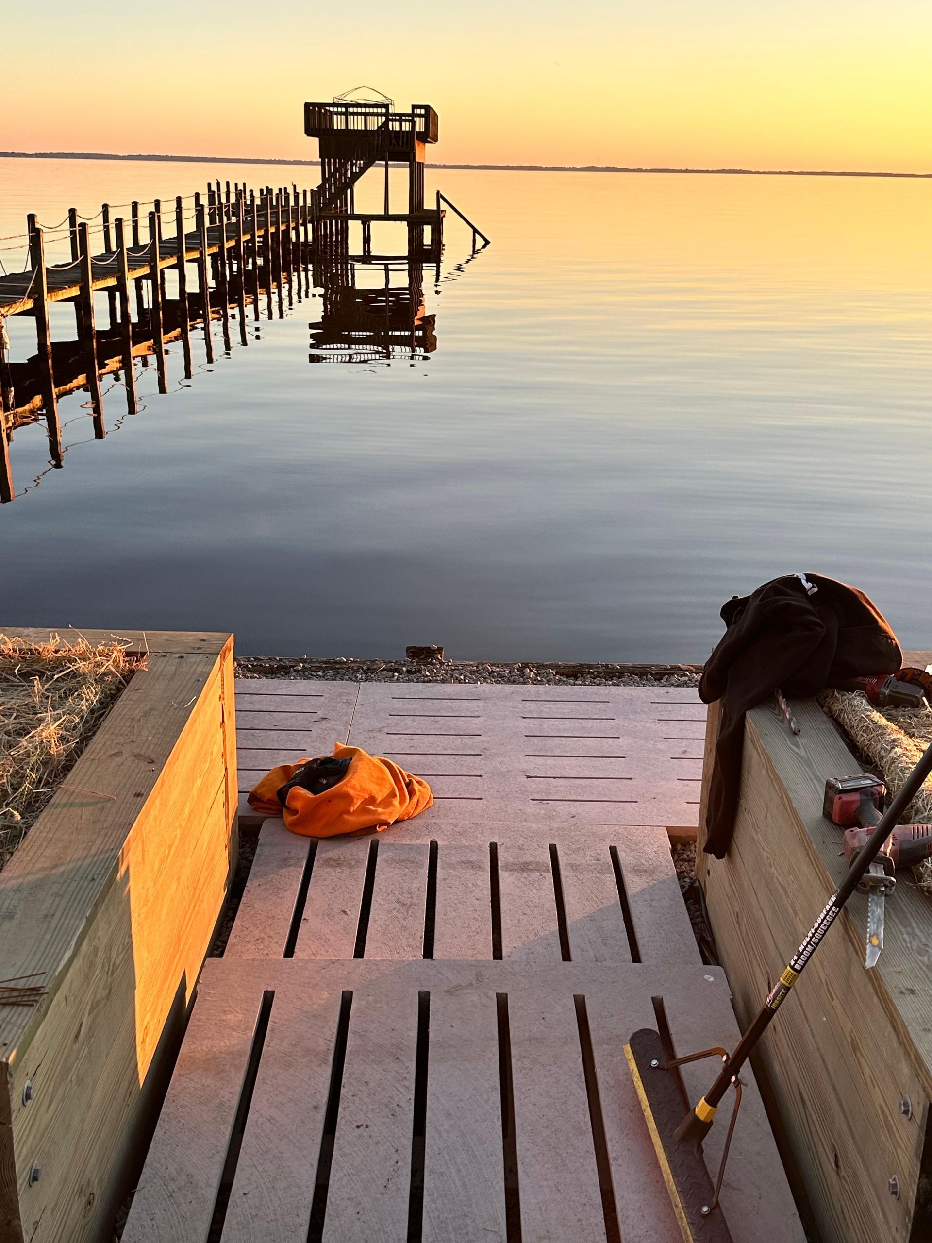 Dockside view: wooden dock, calm water at sunset, orange life jacket, and a push broom.
