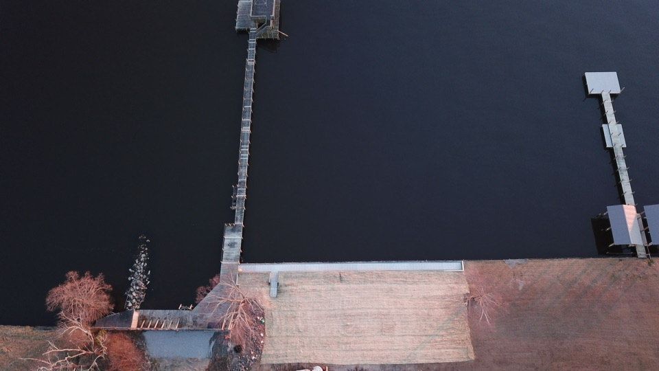 Aerial view of a dock extending into dark water, adjacent to a grassy embankment, at sunset.