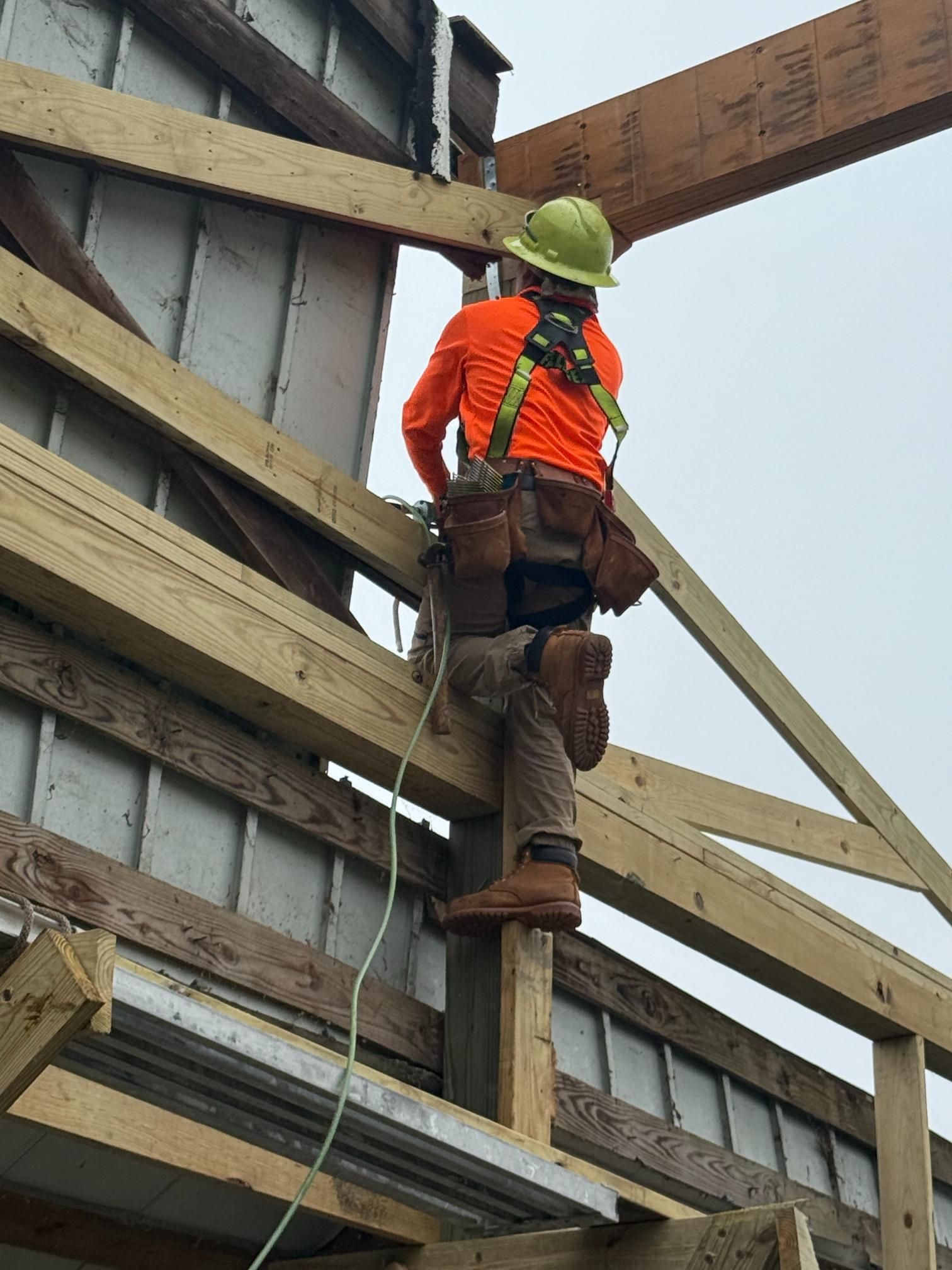 Construction worker in safety gear climbs wooden roof frame on a cloudy day.