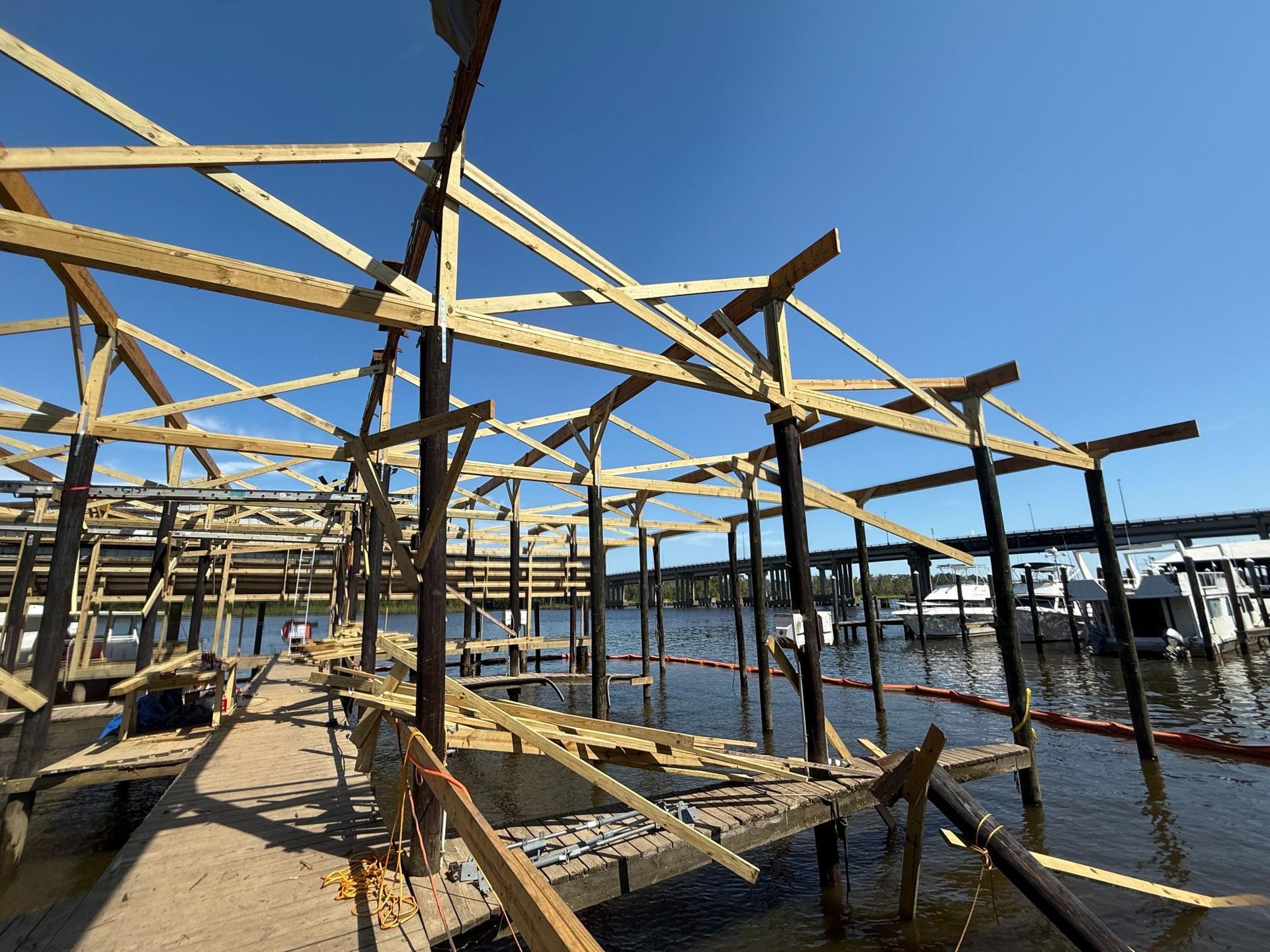 Construction of a wooden boat dock on a waterfront, with support beams, and a bridge in the background.