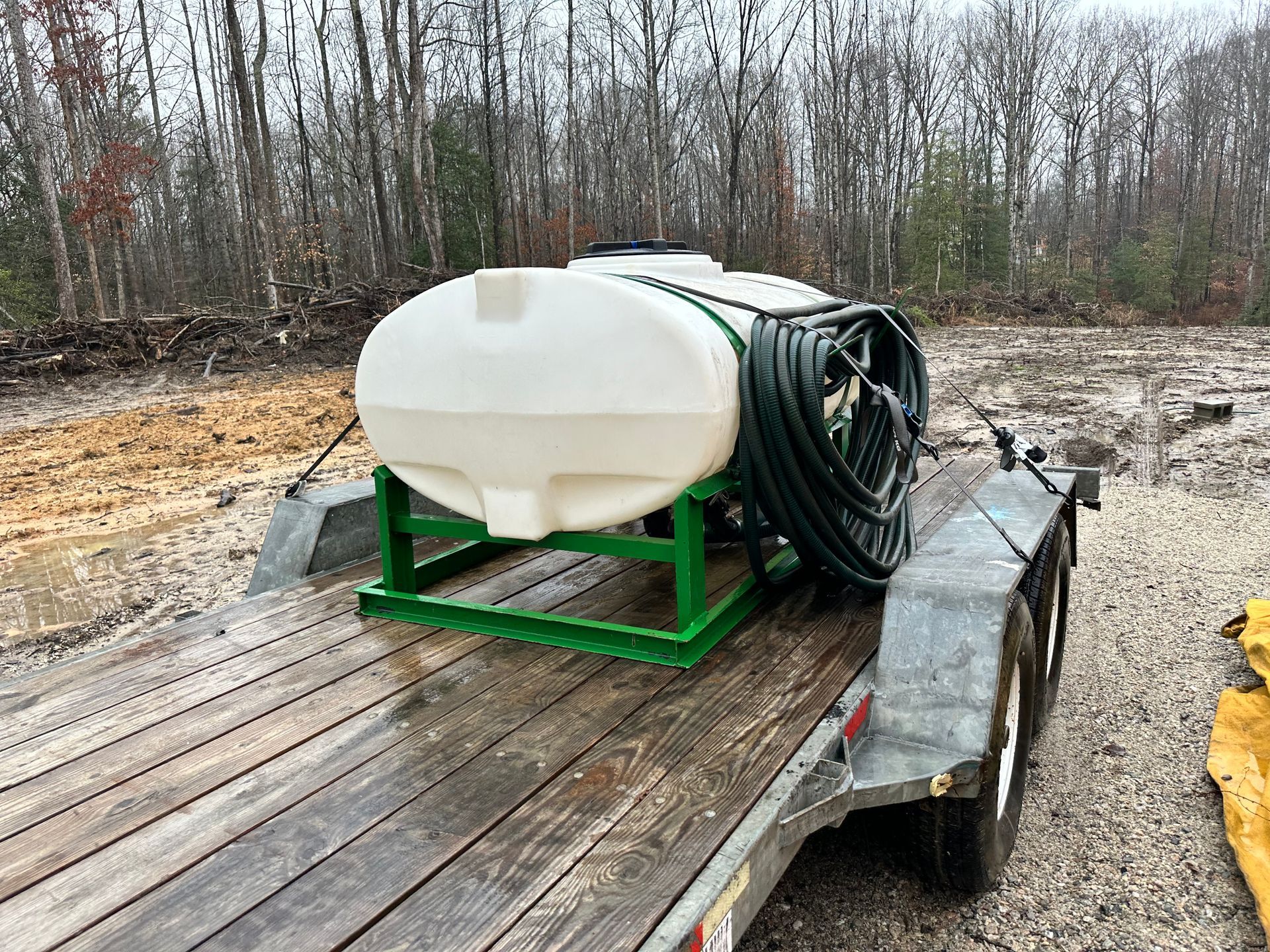 A white water tank on a green metal frame is mounted on a gray trailer, in a muddy outdoor setting.