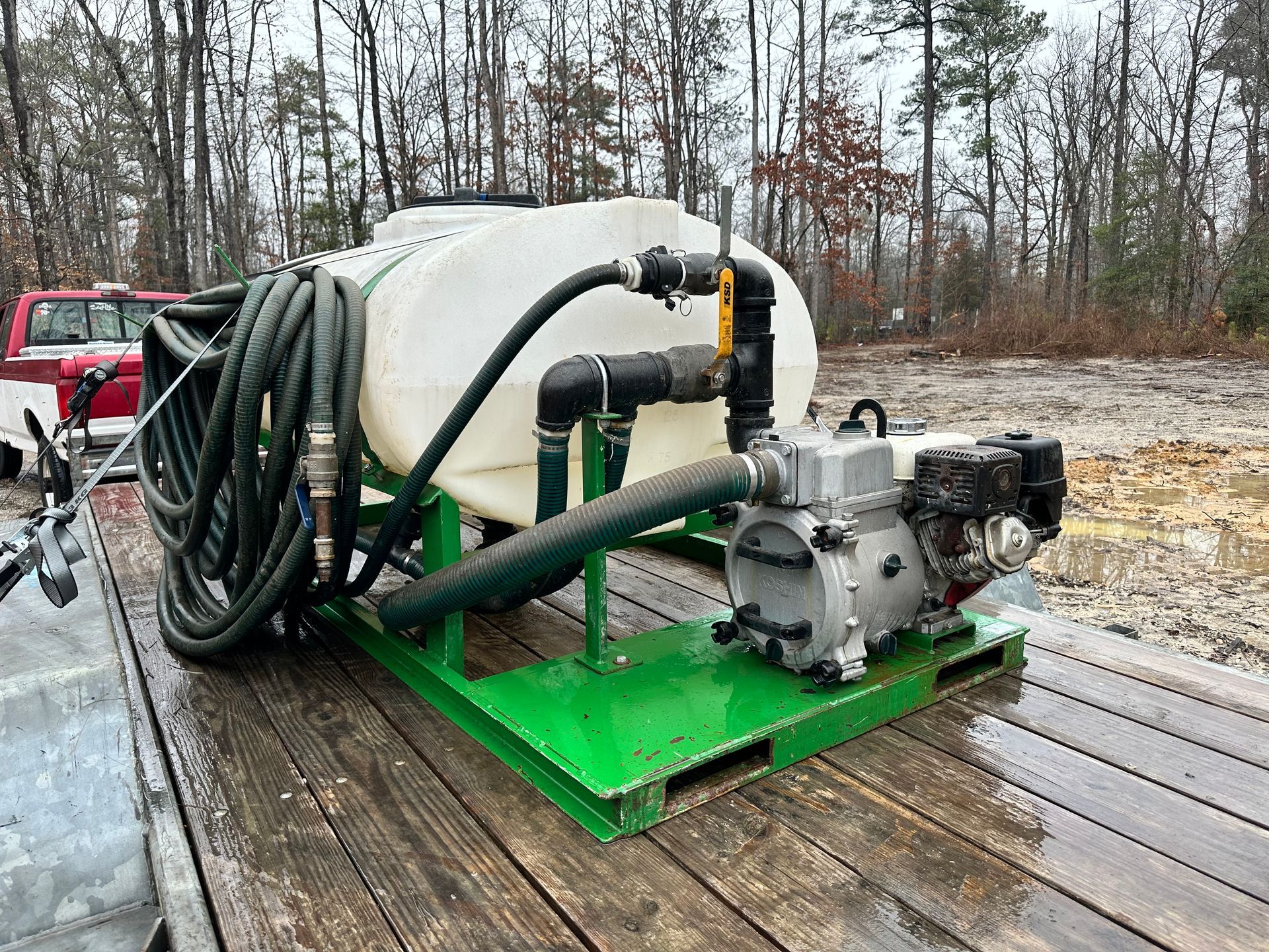 A green pallet holding a white tank, black hoses, pump, and engine on a trailer.