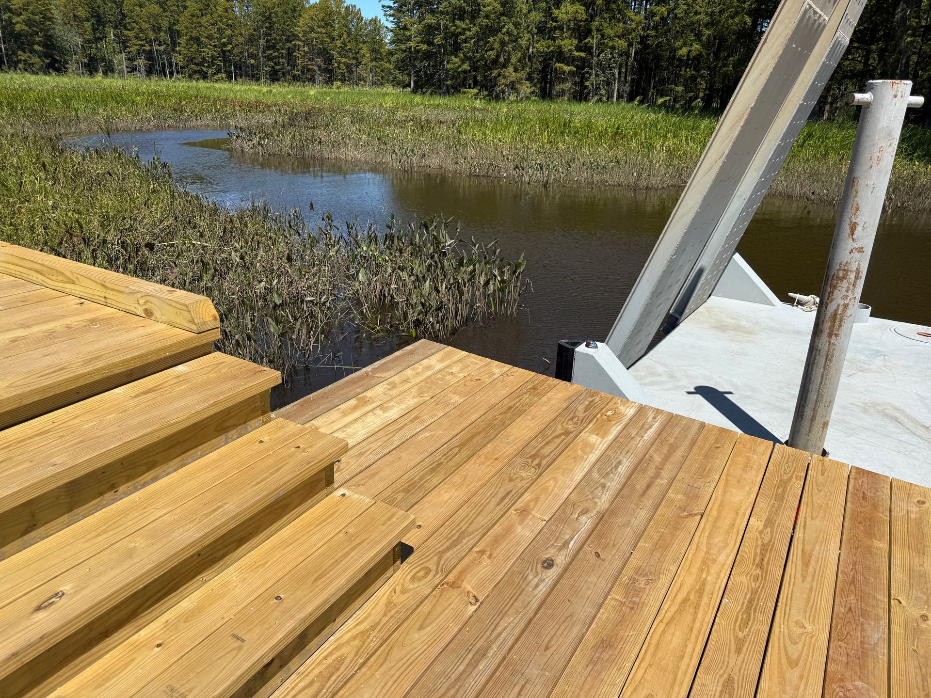 Wooden dock with stairs leading to a body of water and a ramp, reeds in the background.