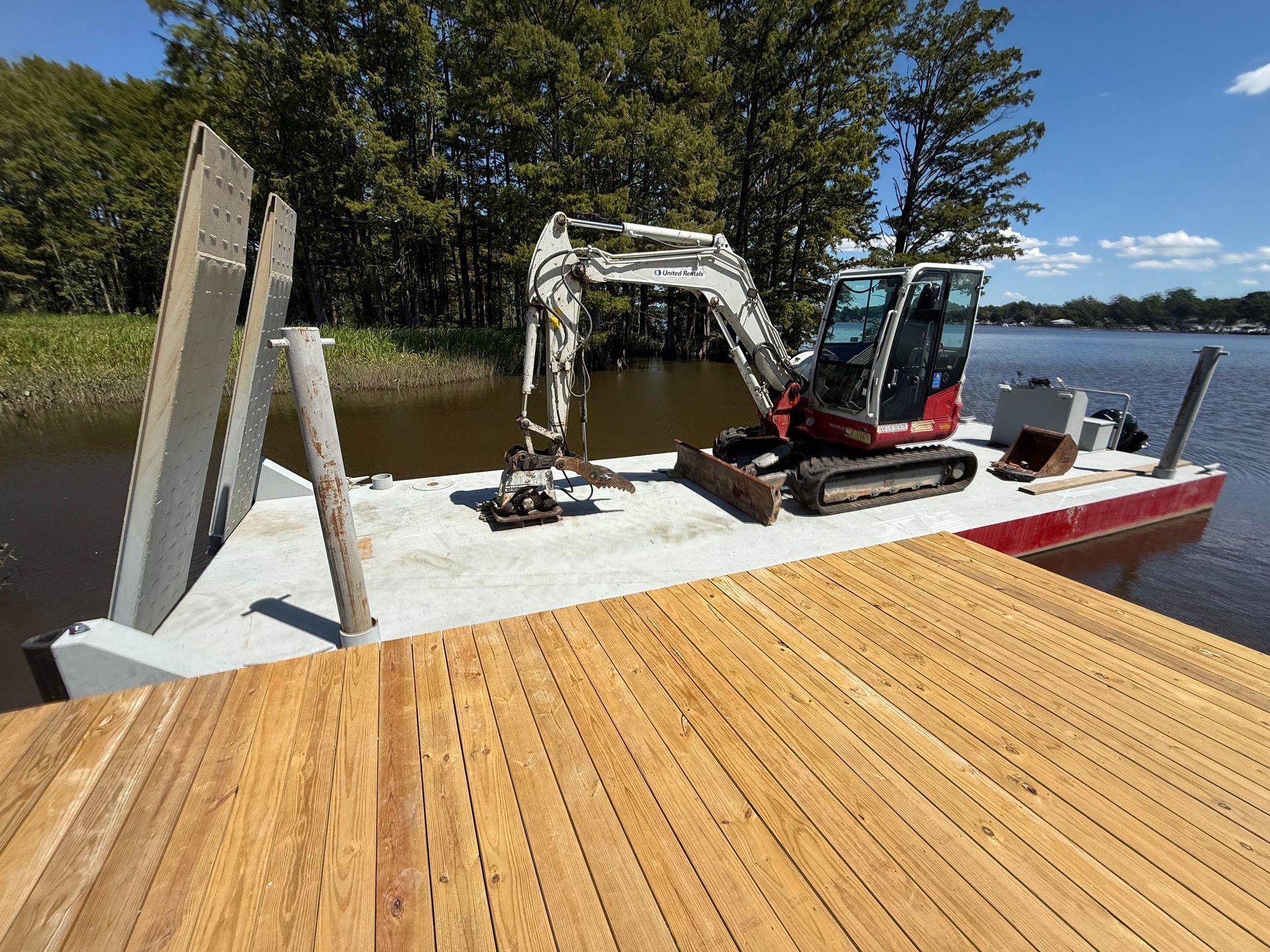 Excavator on a barge, working on a wooden dock at a lake.