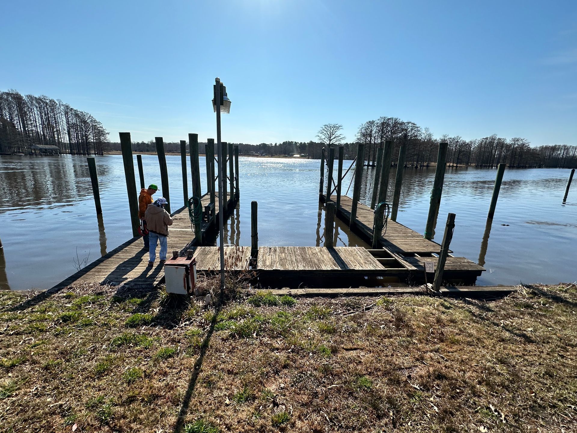 Two people stand on a dilapidated wooden dock extending into a sunlit body of water.