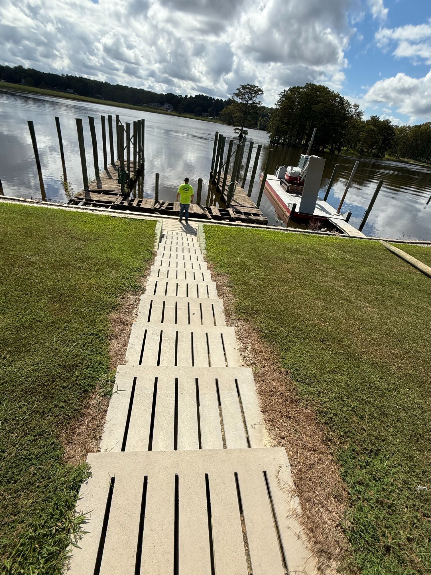 Concrete steps lead down to a dock with a person in a bright vest, near water and green grass.