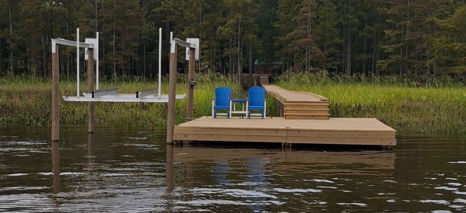 A wooden dock with two blue chairs and a lift in the water, in front of a forest.