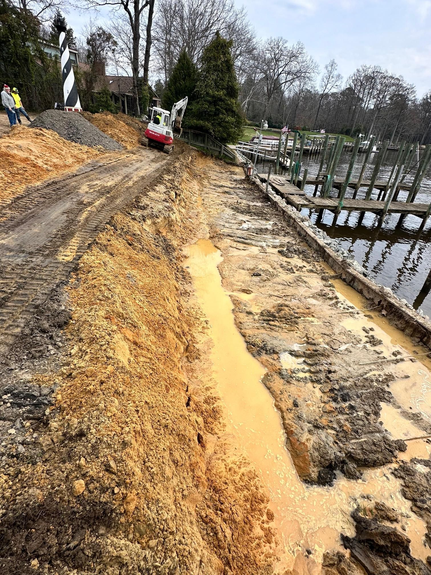 Construction site next to a dock. An excavator digs along the water's edge, earth is muddy and brown.