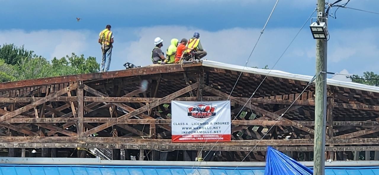 Construction workers on a weathered roof. A banner hangs below the roof's structure. Blue and green hues in the surroundings.