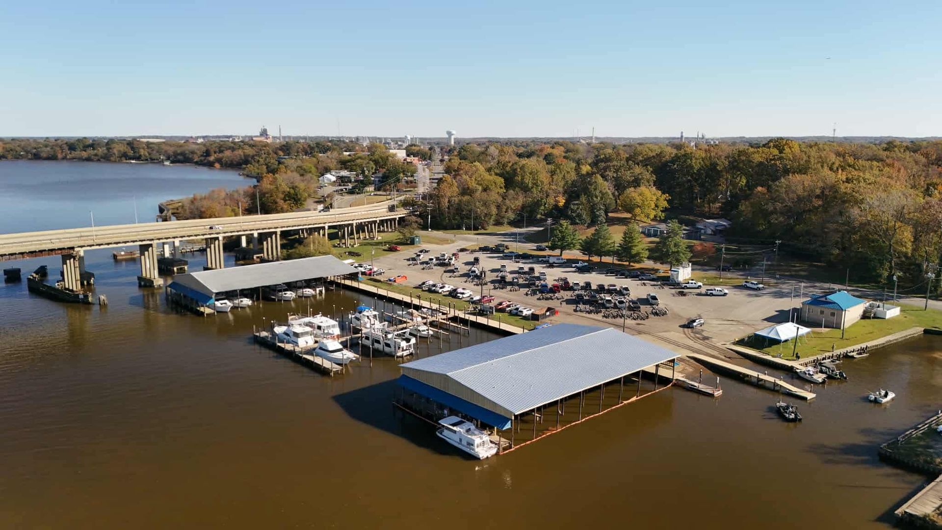 Marina with boats, docks, and a bridge under a clear, sunny sky with trees along the shore.