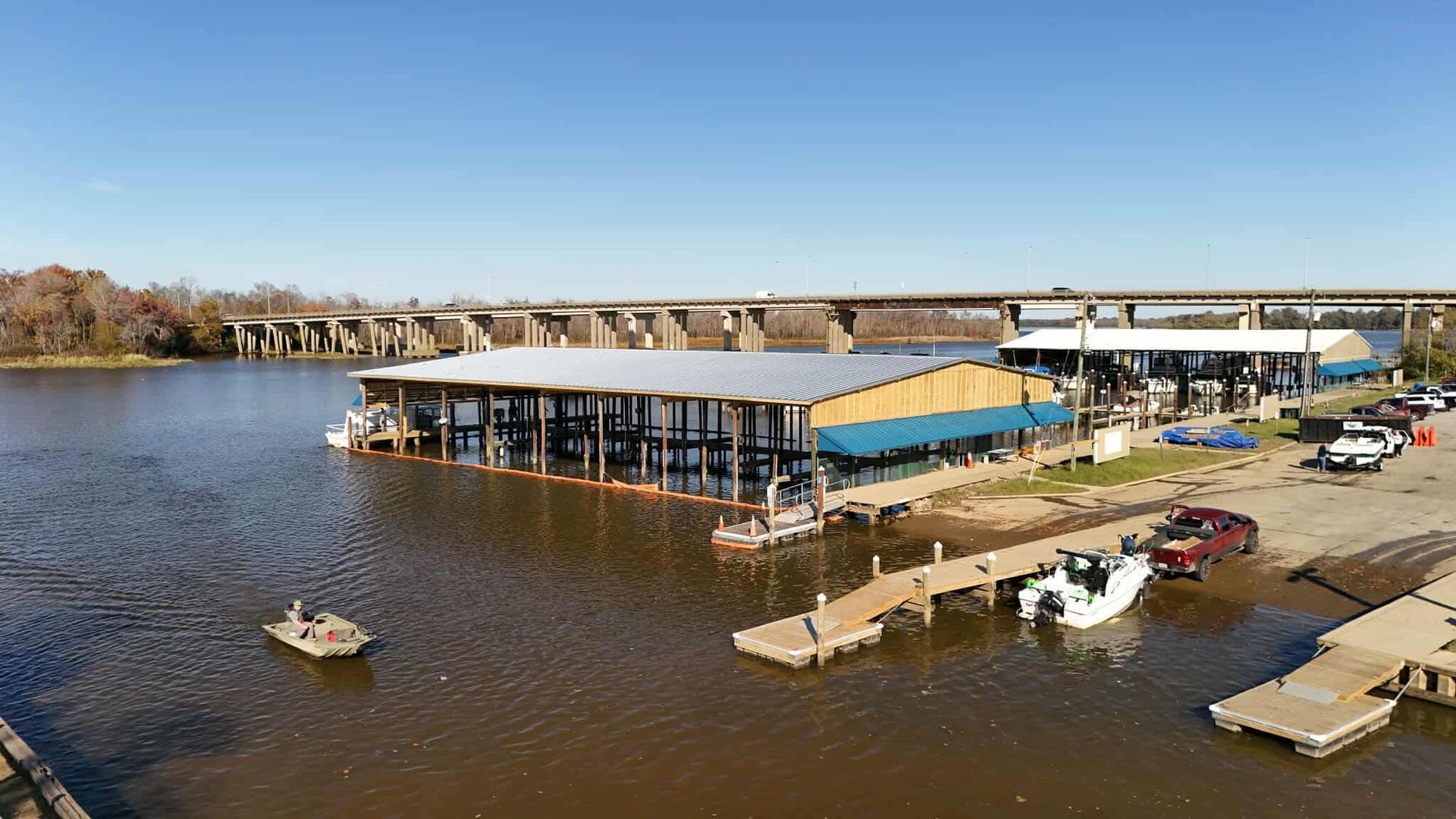 Boathouse on river with bridge in background; boats docked, cars parked. Blue sky.