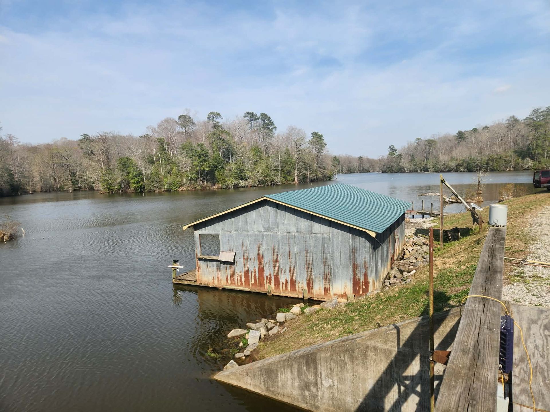 A rustic, tin-sided boathouse with a green metal roof sits on the edge of a calm, tree-lined lake under a blue sky.