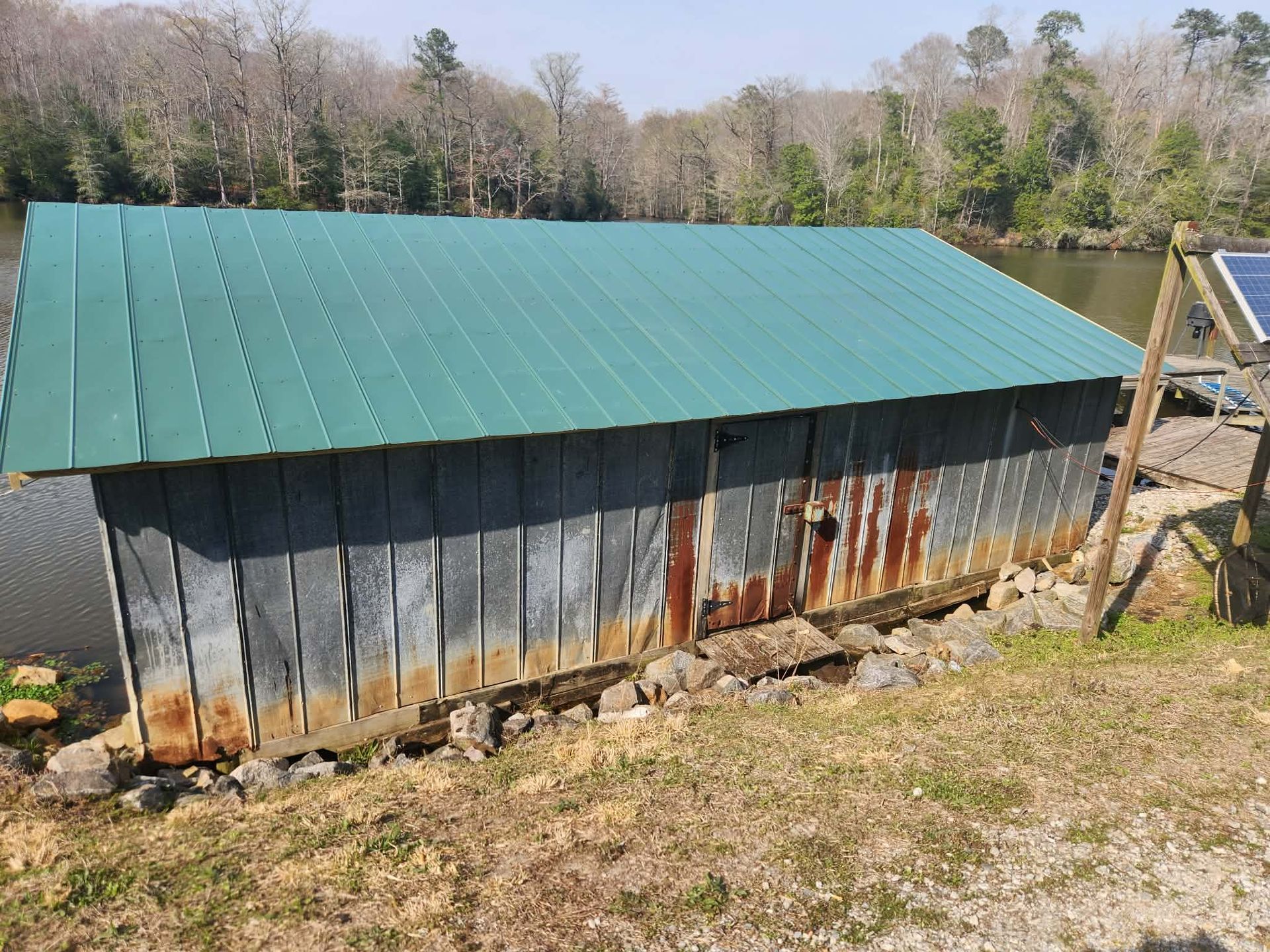A rustic shed with a green metal roof and weathered, rusted corrugated walls sits near a calm lake on a sunny day.