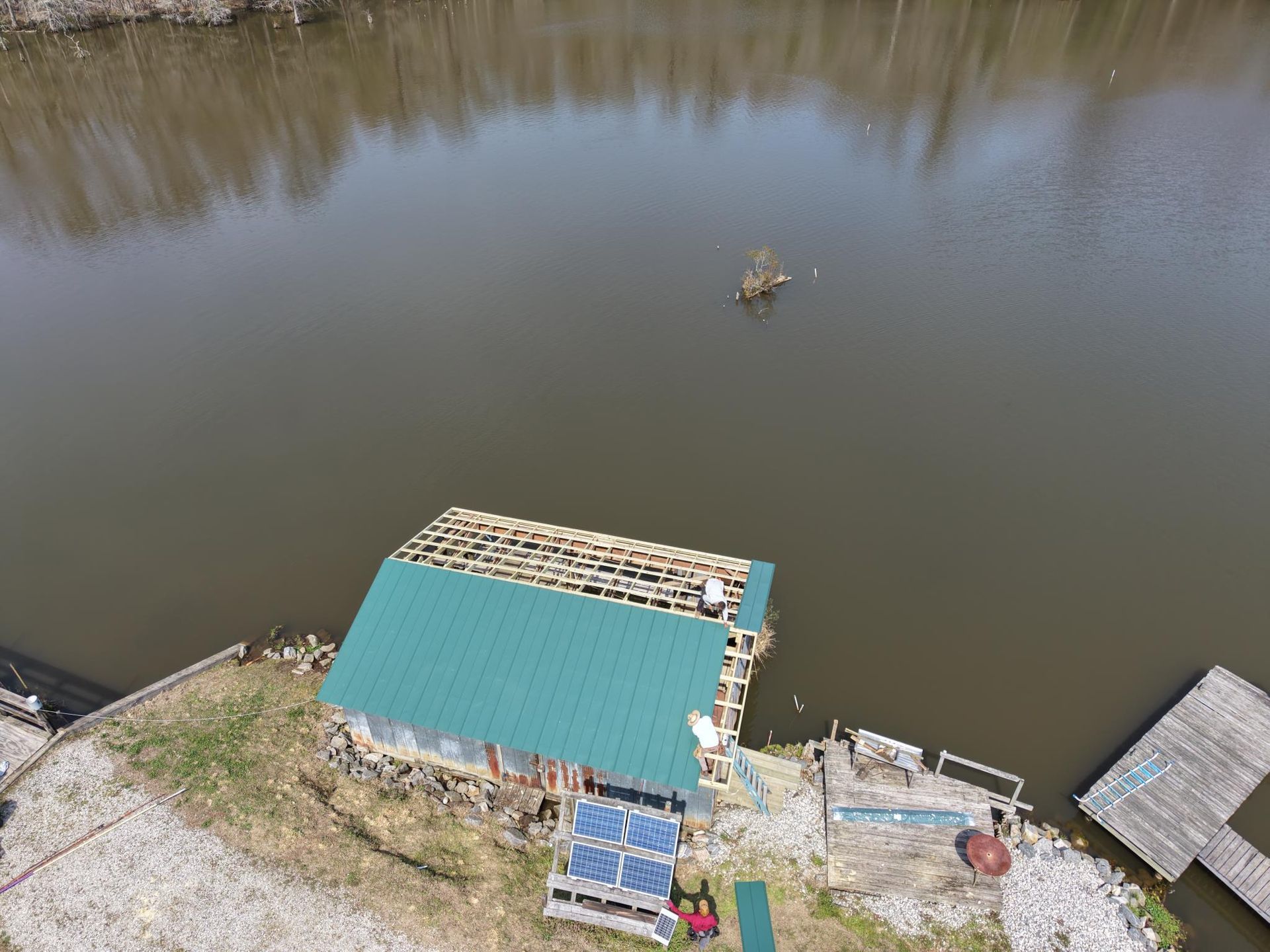 An aerial view of a building with a half-installed green metal roof and exposed rafters by a lake with a small dock.