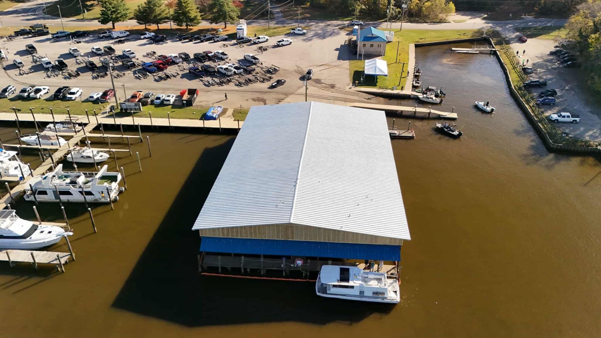 Boats docked at a marina; white roofed structure on water; cars parked in background.