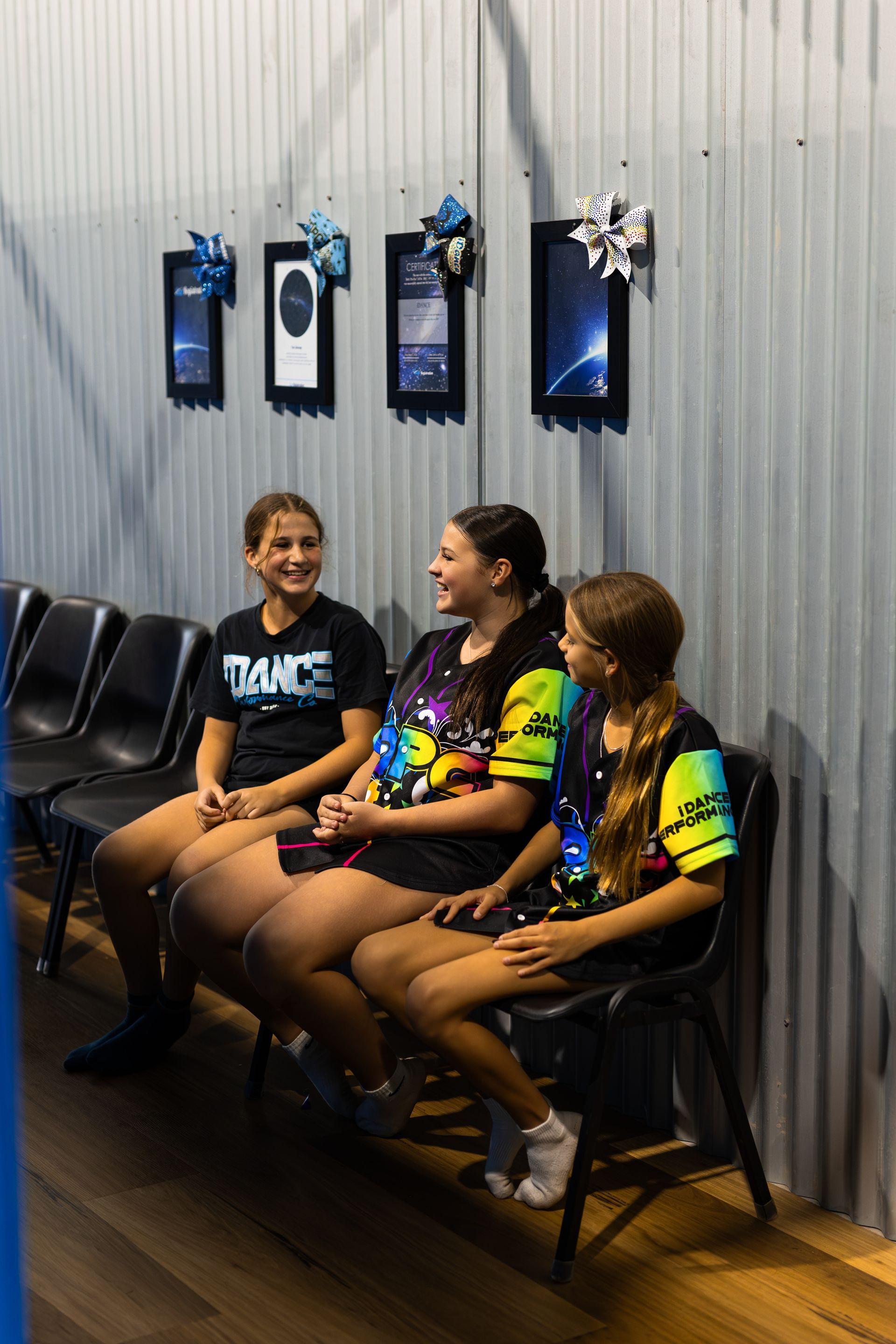 Three Girls in Dance Attire Seated, Smiling. They Are in a Room With Framed Displays — iDance Performance Co In Medowie, NSW