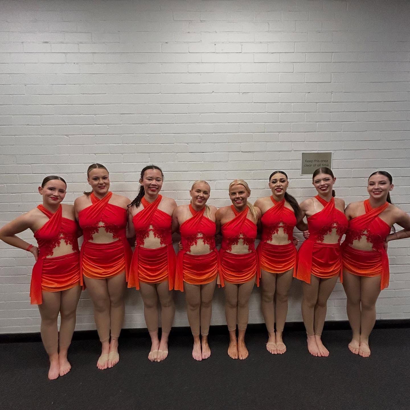 Seven Dancers in Orange and Red Costumes Stand in a Row Against a White Brick Wall — iDance Performance Co In Medowie, NSW