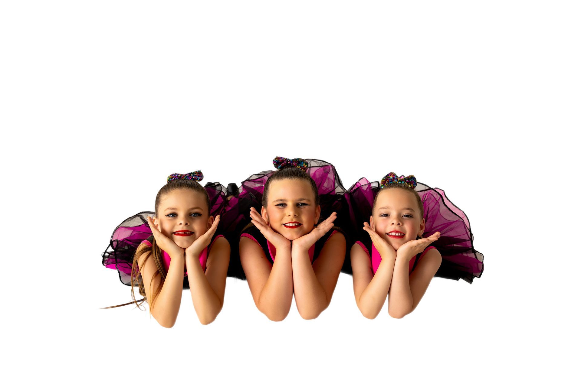 Three young dancers in pink tops and purple tutus, smiling with hands framing their faces — iDance Performance Co In Medowie, NSW