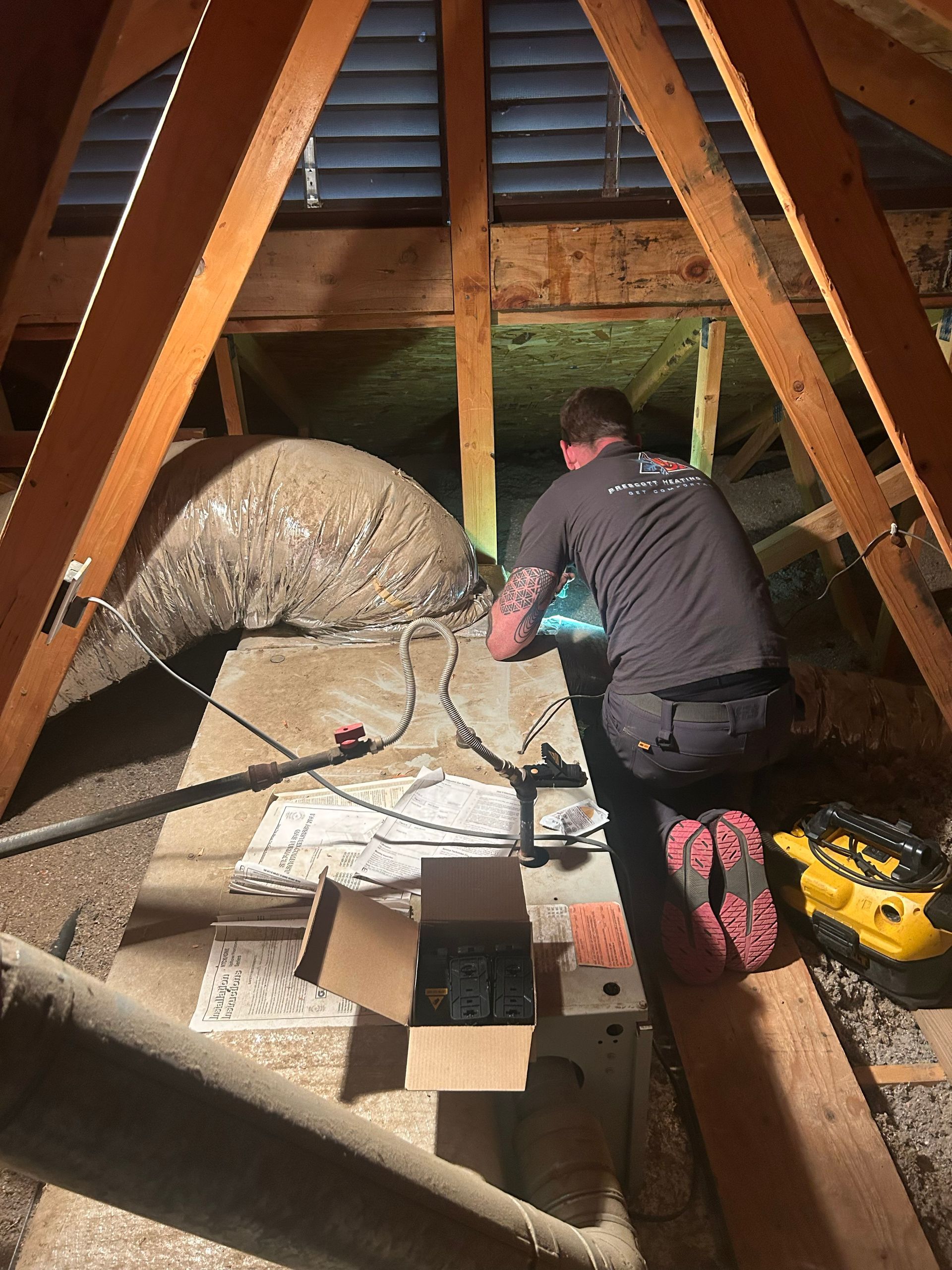 Man in attic working on electrical wiring; surrounded by insulation and wooden beams.