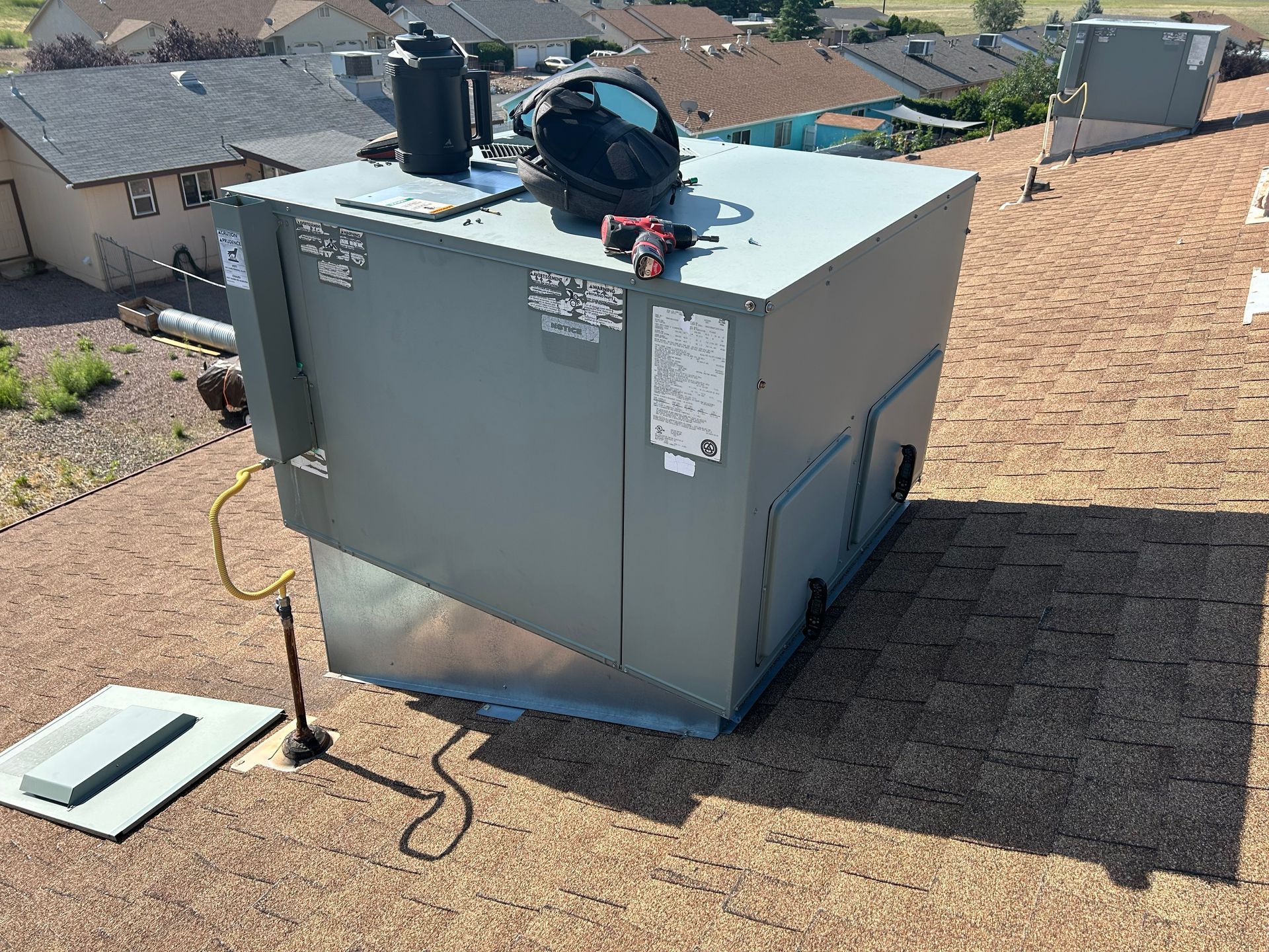 Rooftop HVAC unit, gray, with tools, on a brown shingle roof, surrounded by houses.