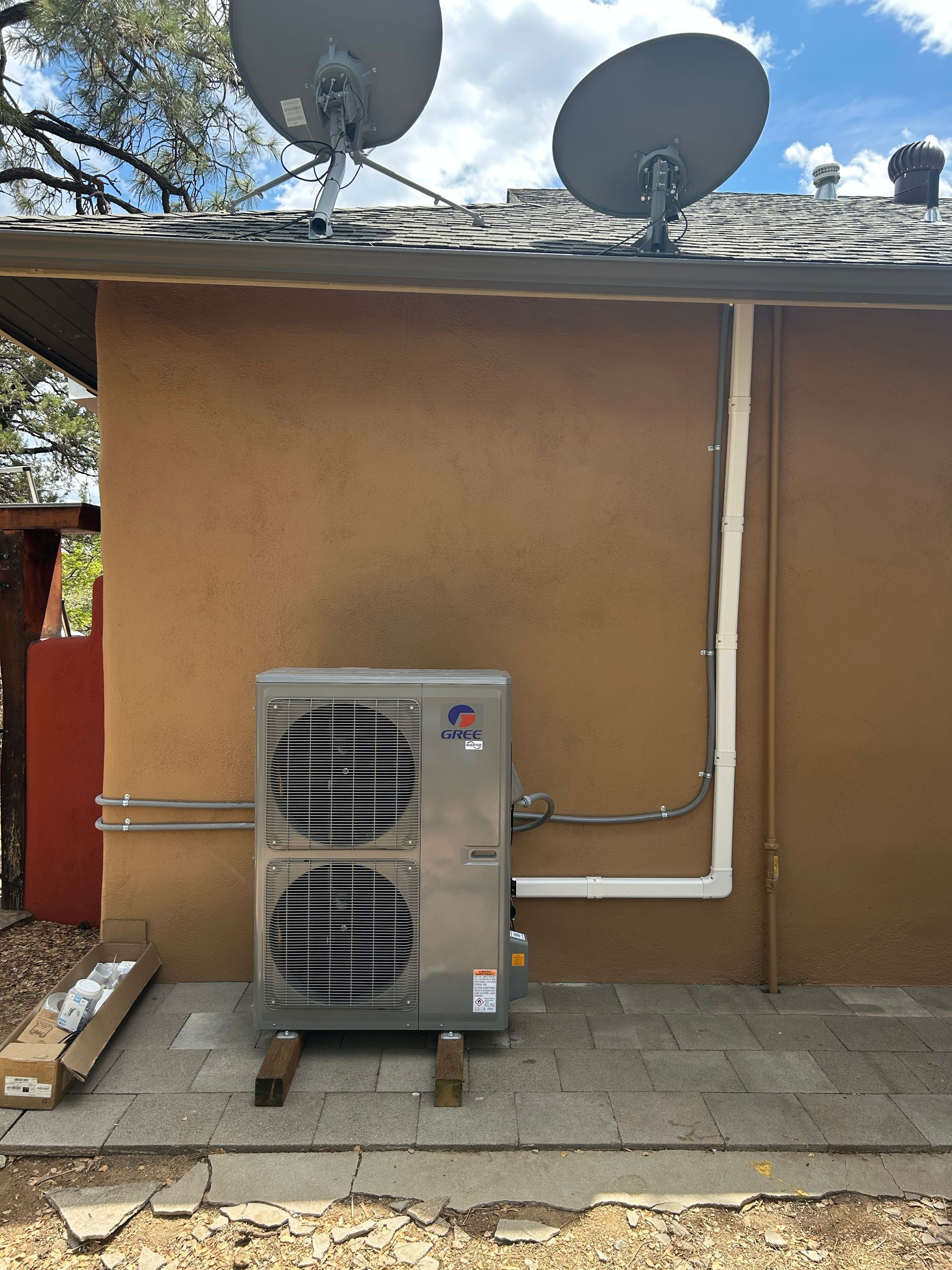 Outdoor air conditioning unit mounted on a brown stucco wall, with satellite dishes above.