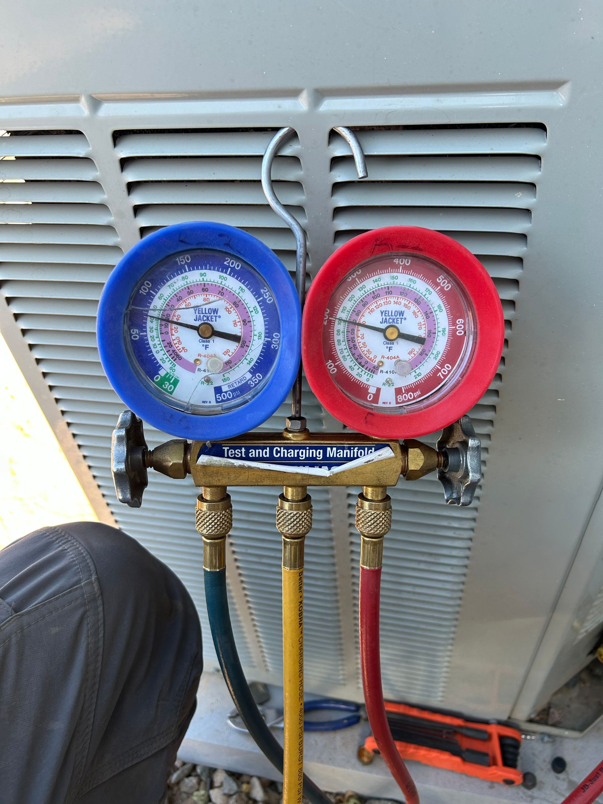 Refrigeration gauge manifold connected to an AC unit, showing pressure levels, with blue and red gauges.