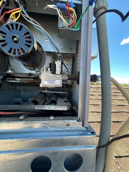 Furnace interior showing gas valve, burner tubes, and blower, on a rooftop with blue sky.