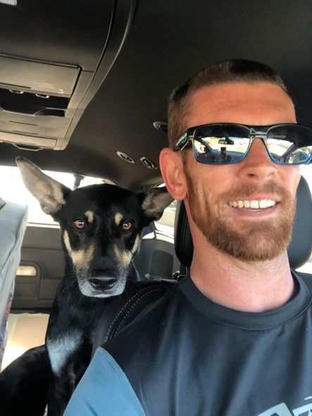 Man with sunglasses smiles at the camera while a black and tan dog sits on his shoulder in a car.