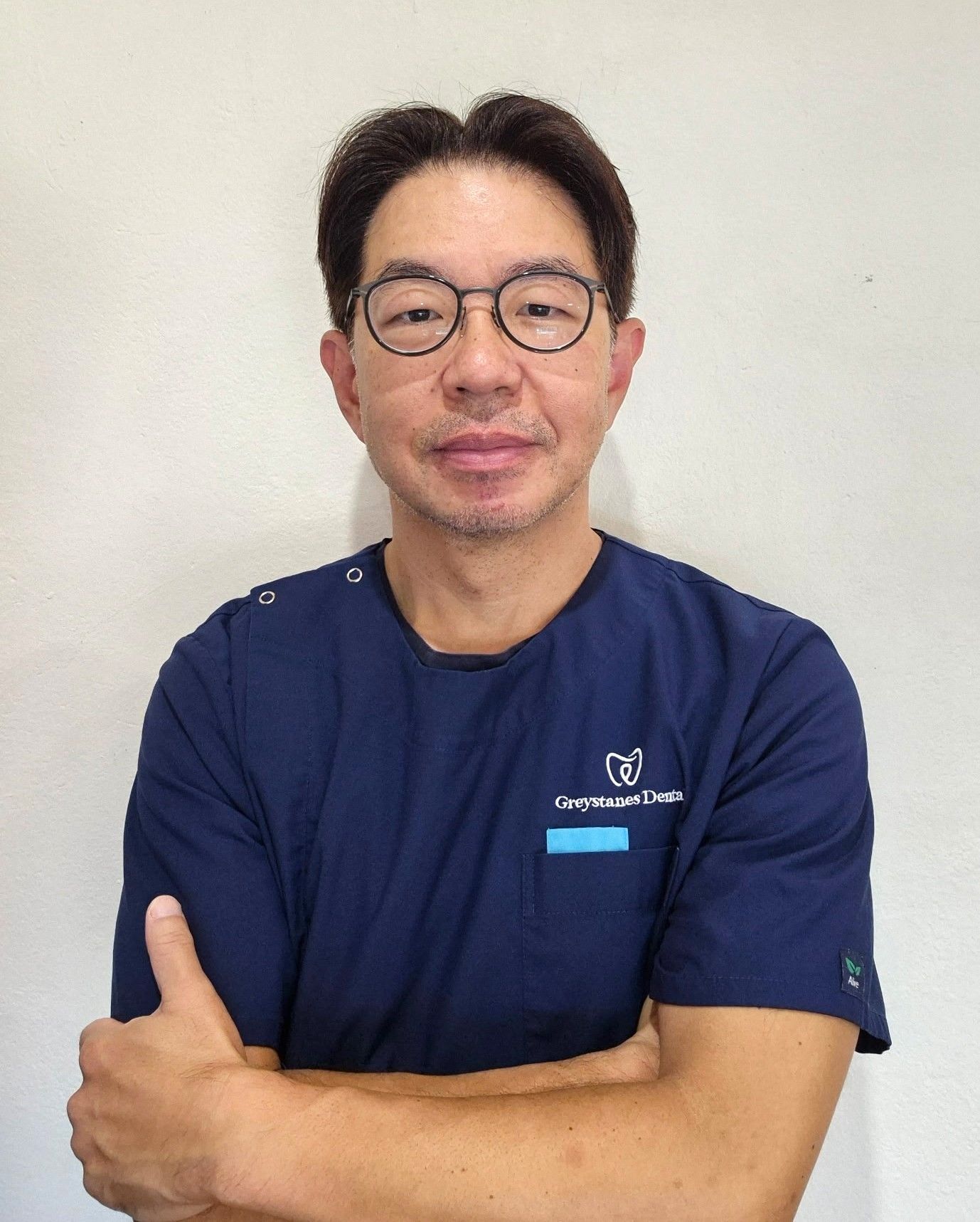 A man wearing glasses and a blue scrub top is standing in front of a white wall.