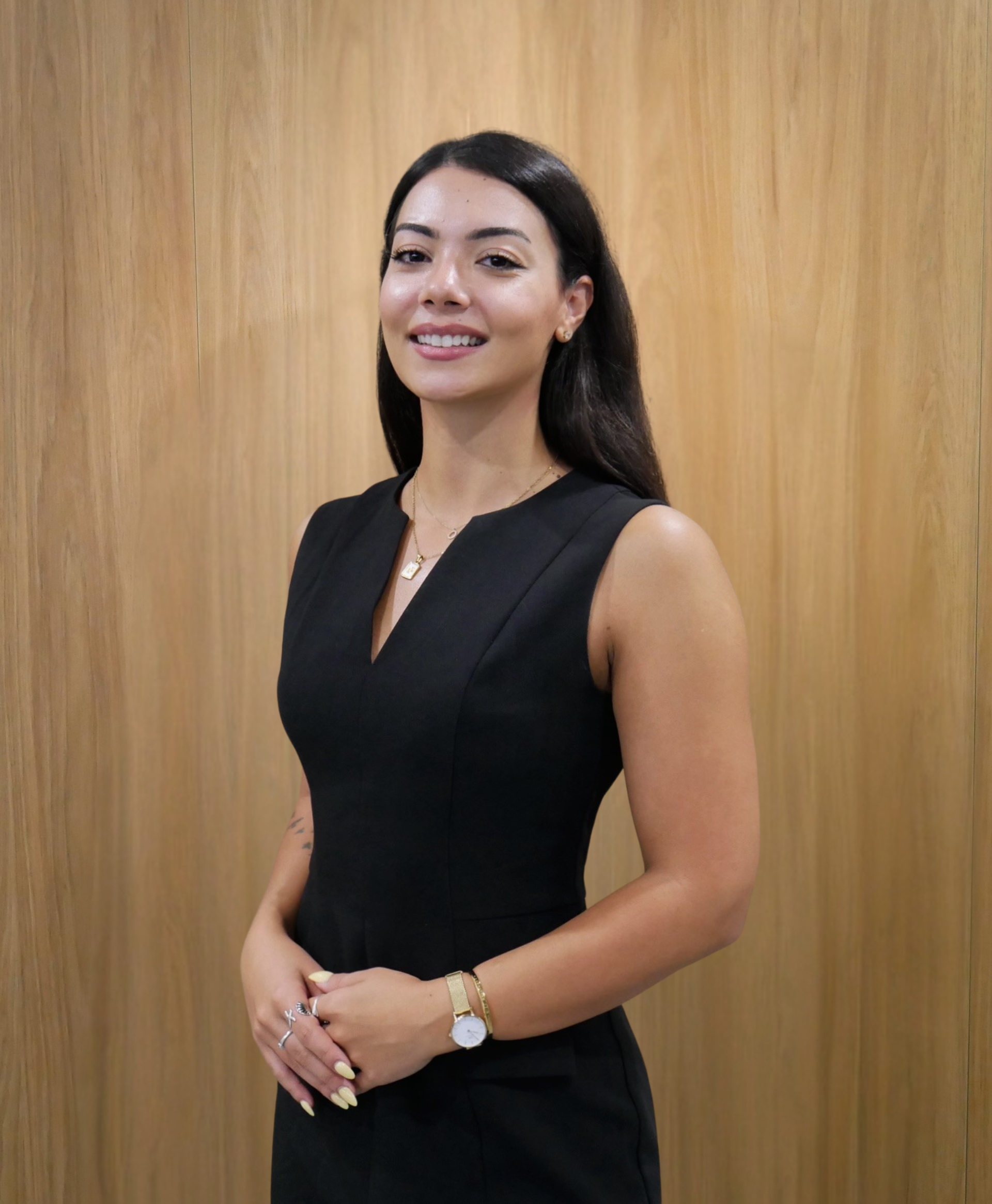 Woman in black sleeveless dress, smiling, arms crossed, wooden background.