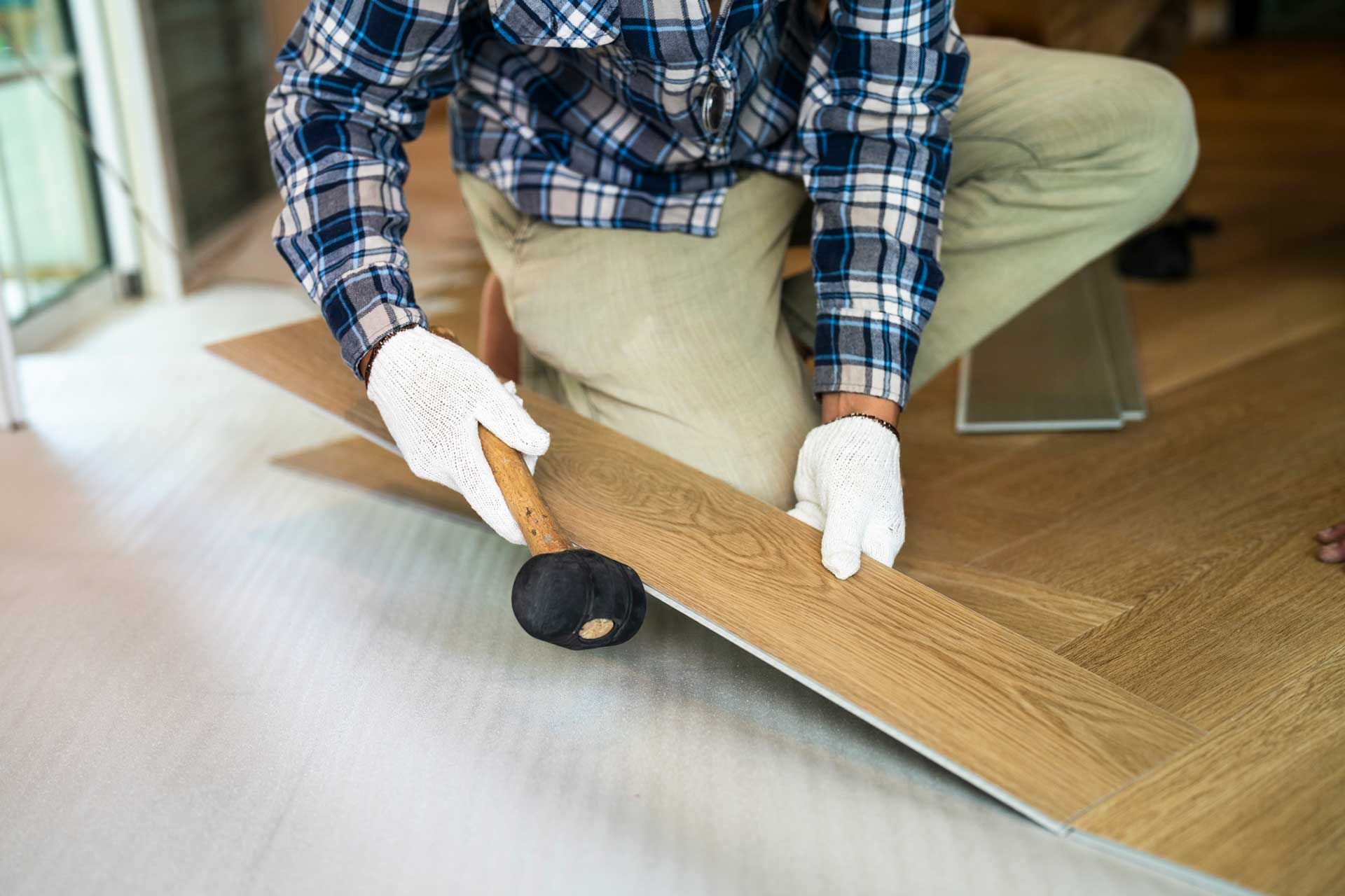 A man wearing gloves holds a hammer while working on hardwood flooring installation in a home.
