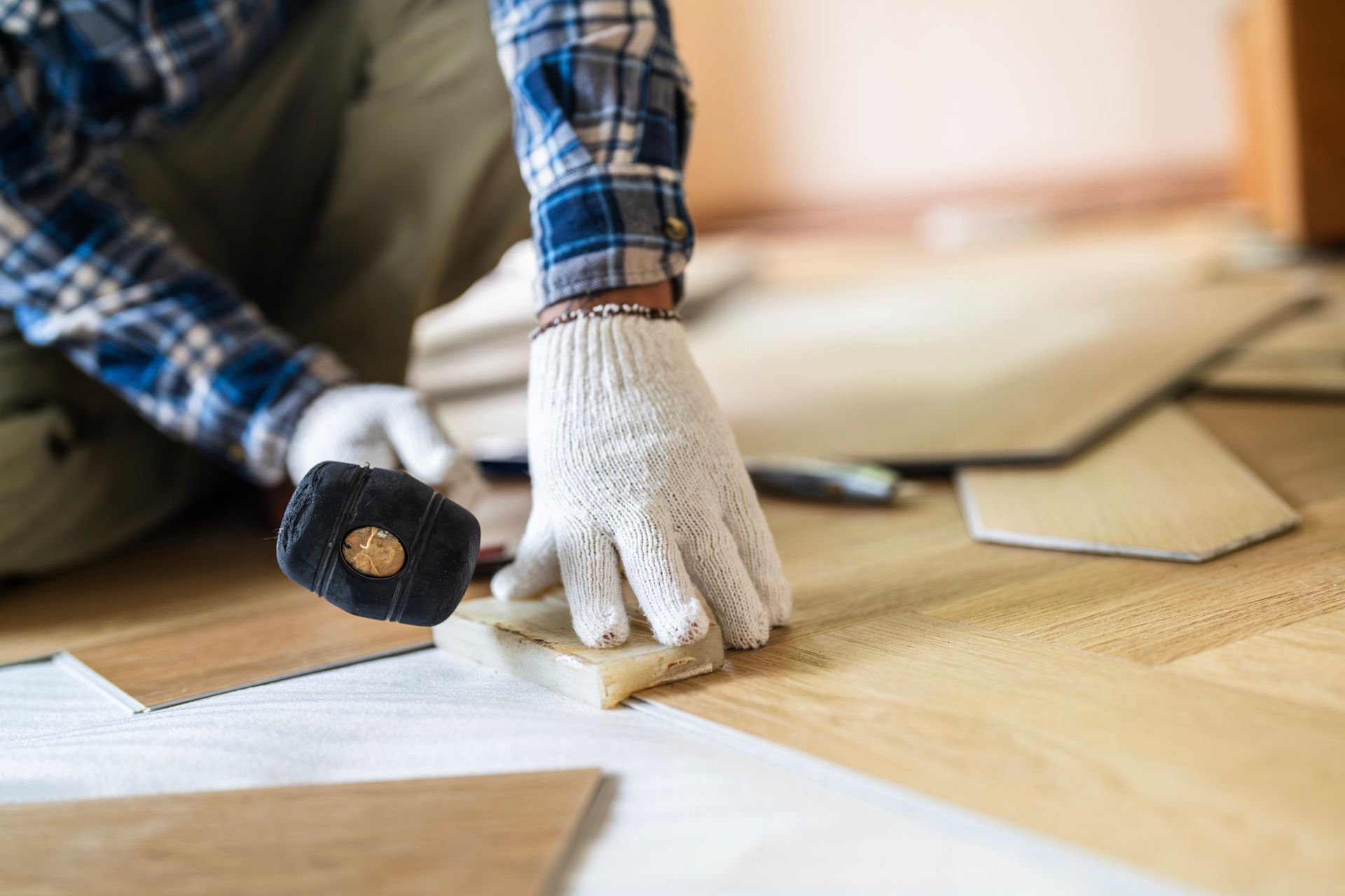A male worker installs laminate flooring with a mallet and gloves.