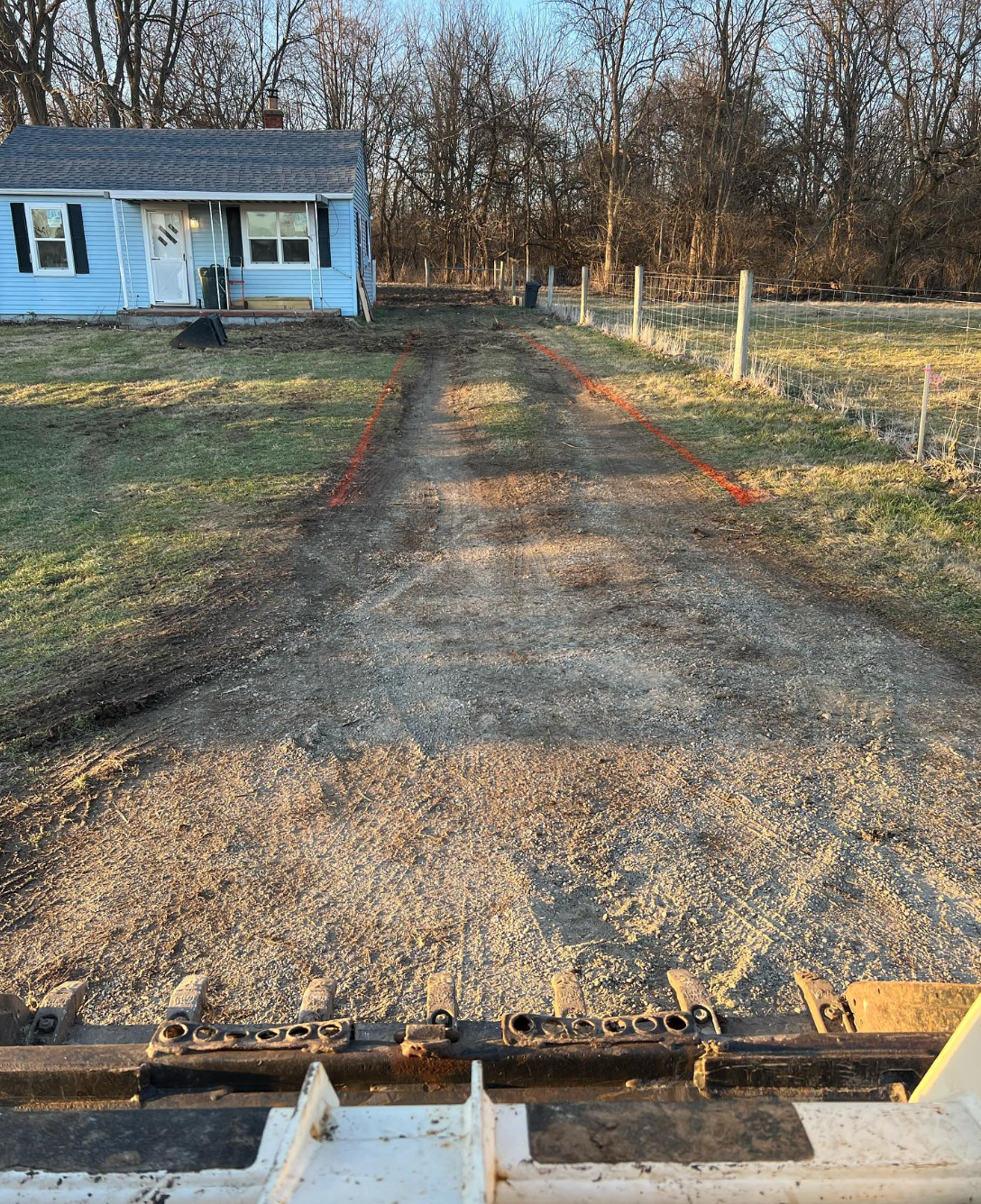 A tractor is driving down a dirt road in front of a house.