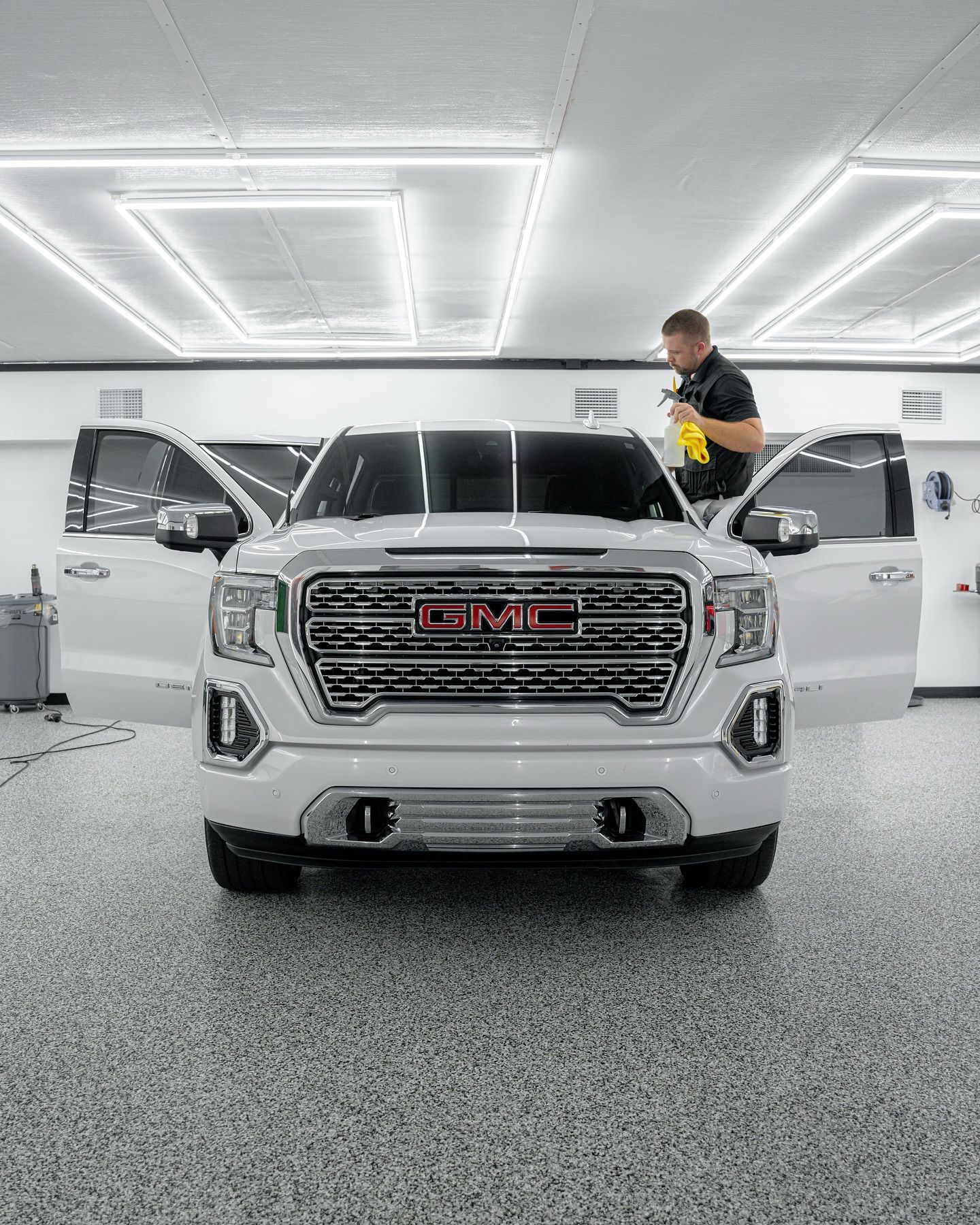 A white GMC truck with open doors, being worked on by a man spraying a window in a well-lit garage.