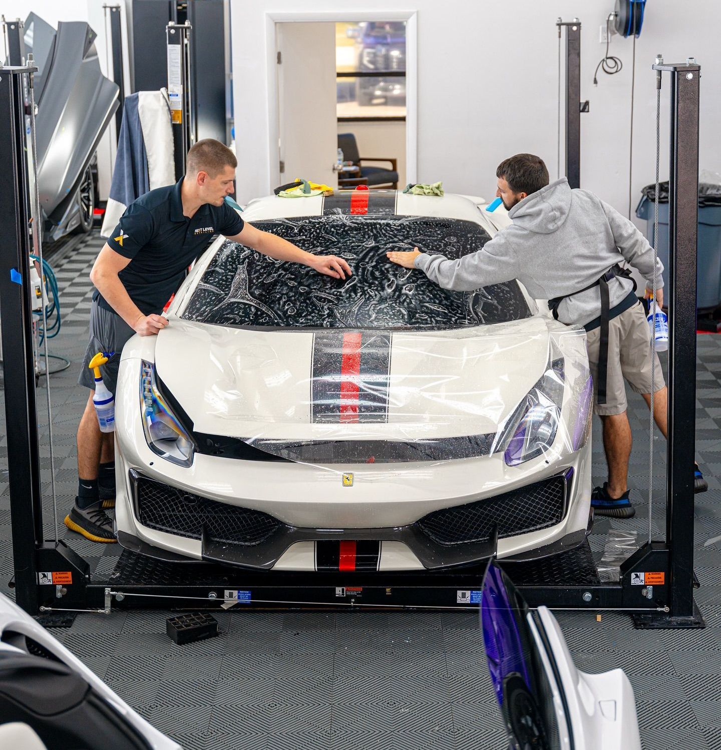 Two men applying clear film to the front of a white Ferrari inside a shop.