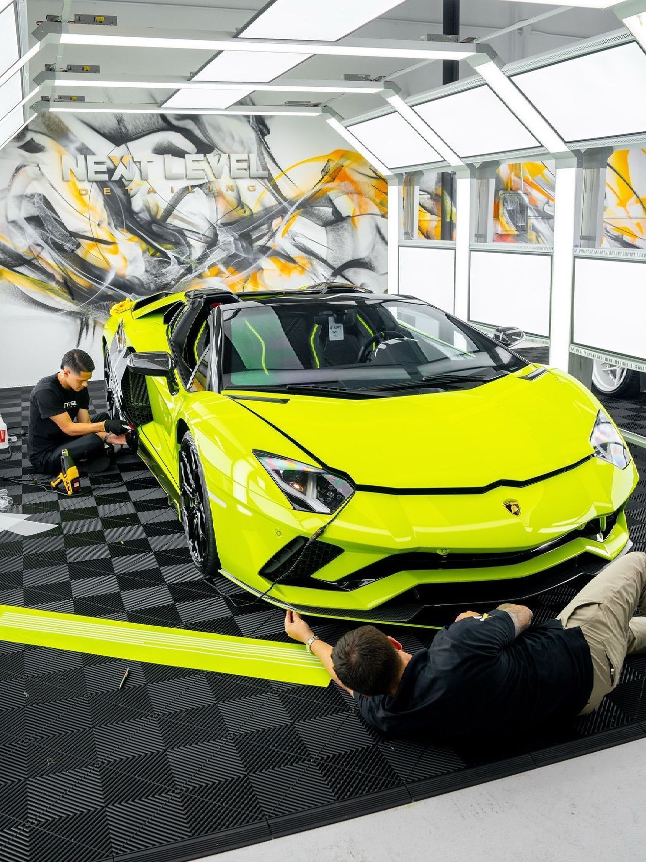 Two men working on a bright yellow Lamborghini in a garage. One is lying down, the other is working on the car's side.
