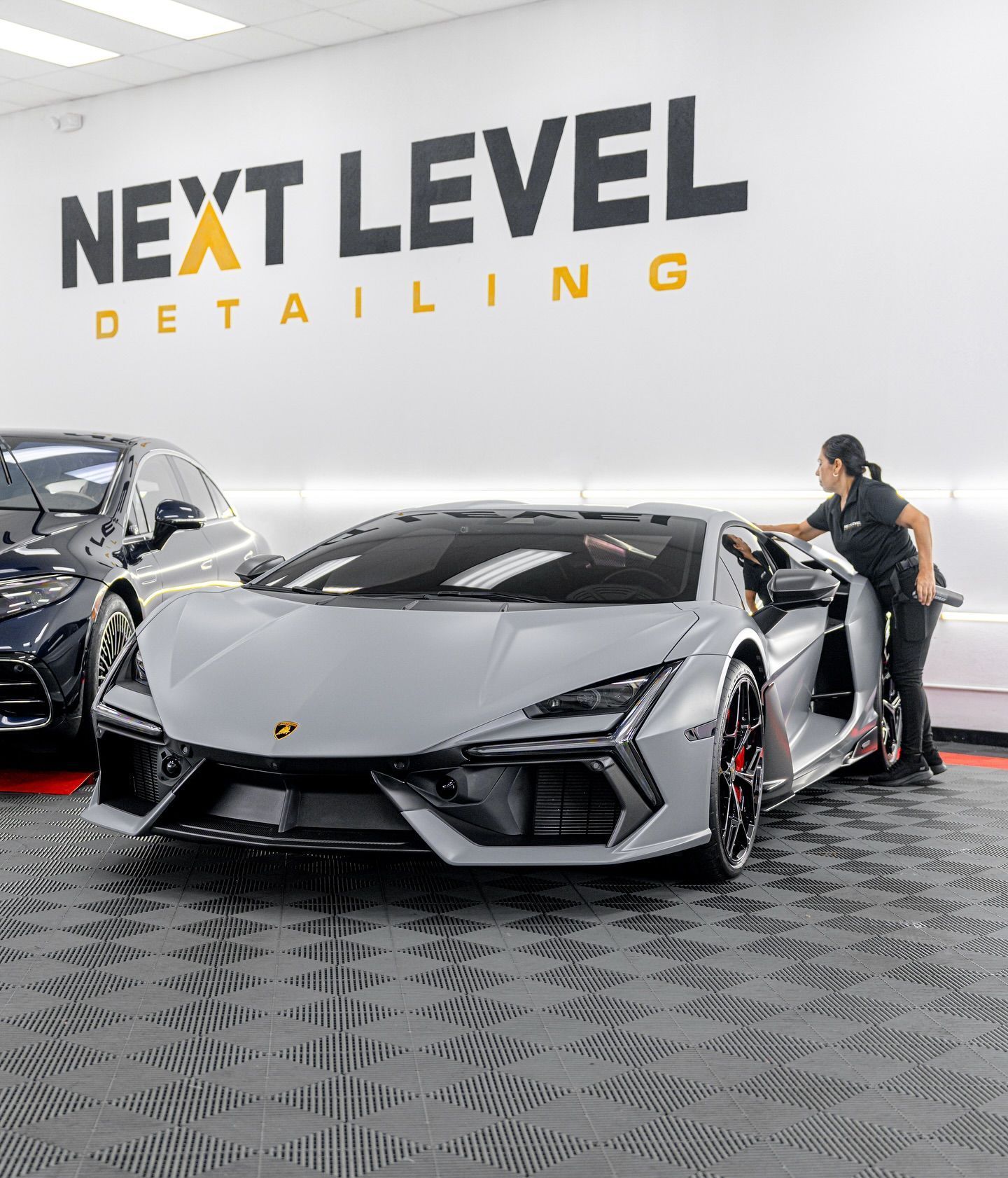 A silver Lamborghini is being detailed inside a shop. A person in a black shirt leans towards the car, while another vehicle sits behind it.