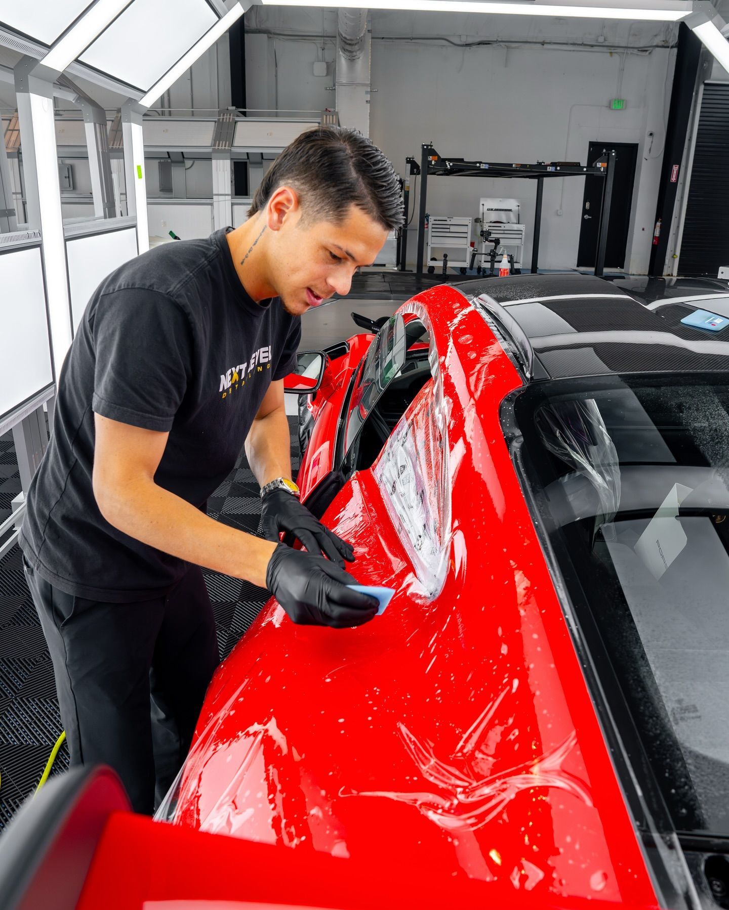 Man in black shirt applying a product to the red hood of a car inside a detailing shop.