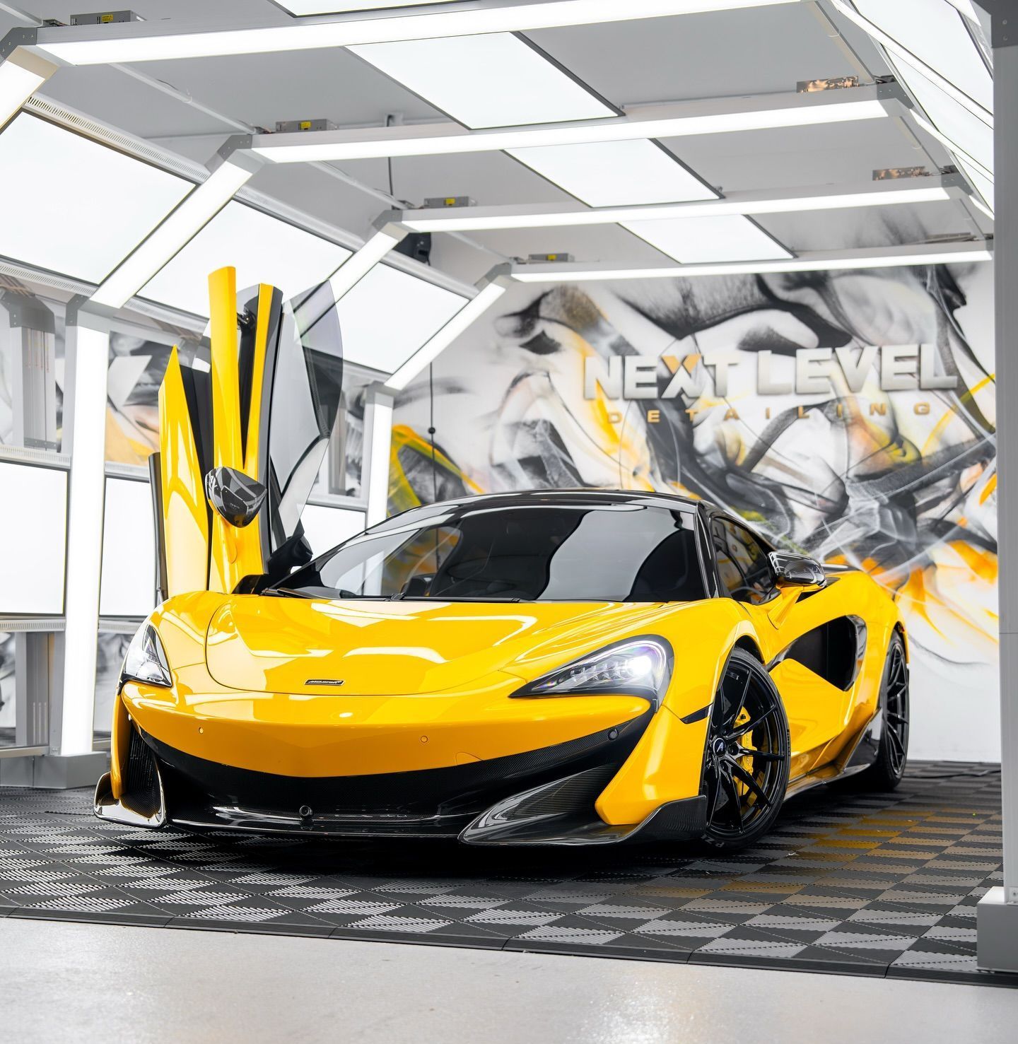 Yellow McLaren sports car with gullwing doors open, parked in a modern garage with 