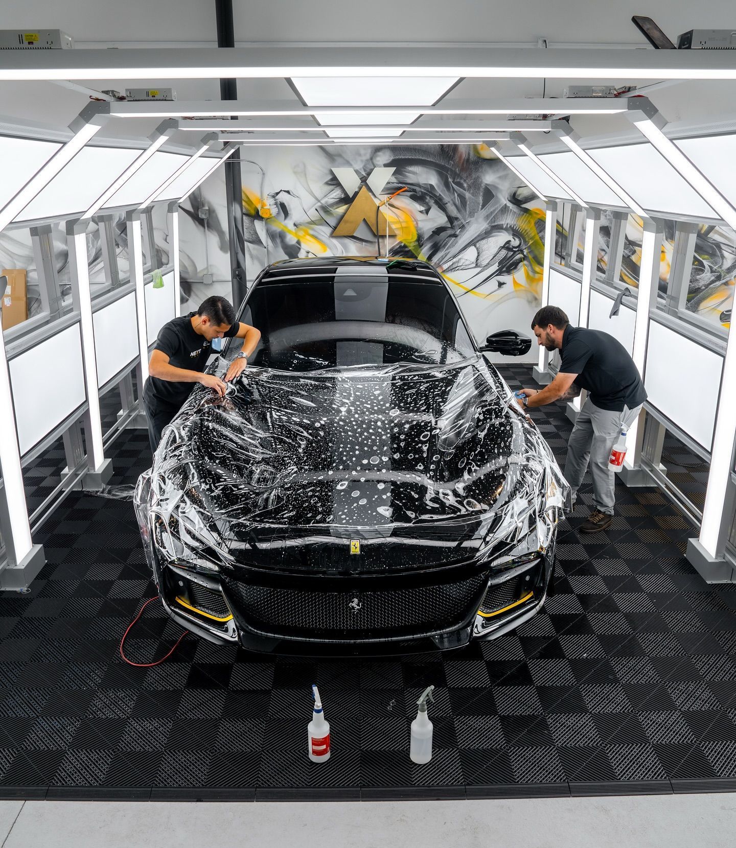 Two people applying protective film to a black car inside a well-lit garage. Spray bottles sit on the floor.