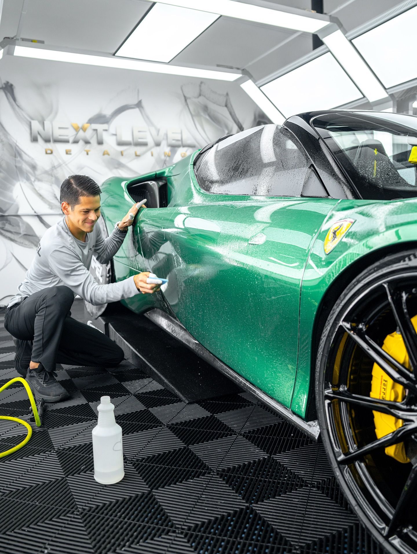 A man applies a film to a green sports car in a detailing shop. He smiles as he works.