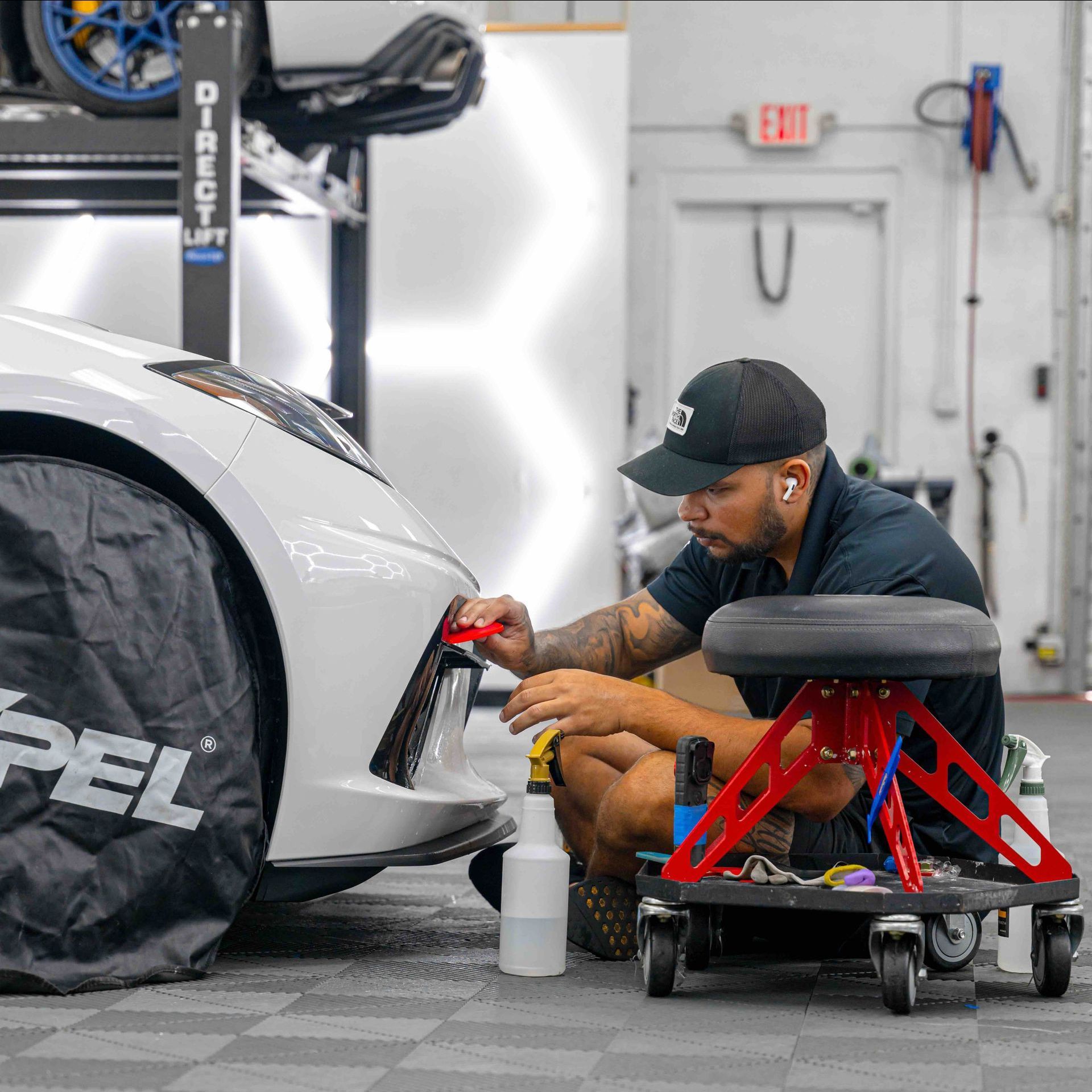 A man is sitting on a stool in front of a car in a garage.