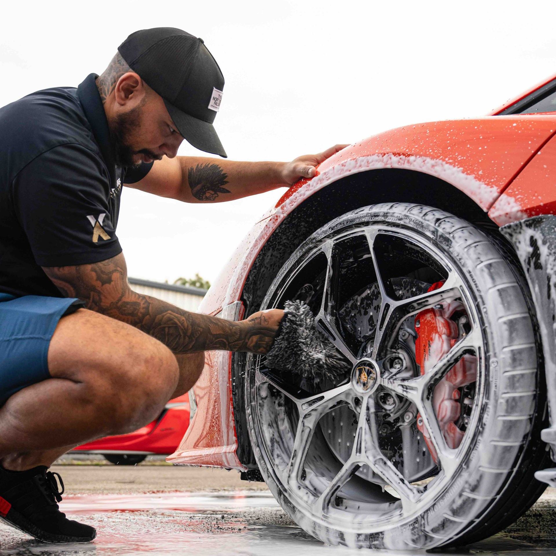 A man is kneeling down to wash the wheels of a red sports car.